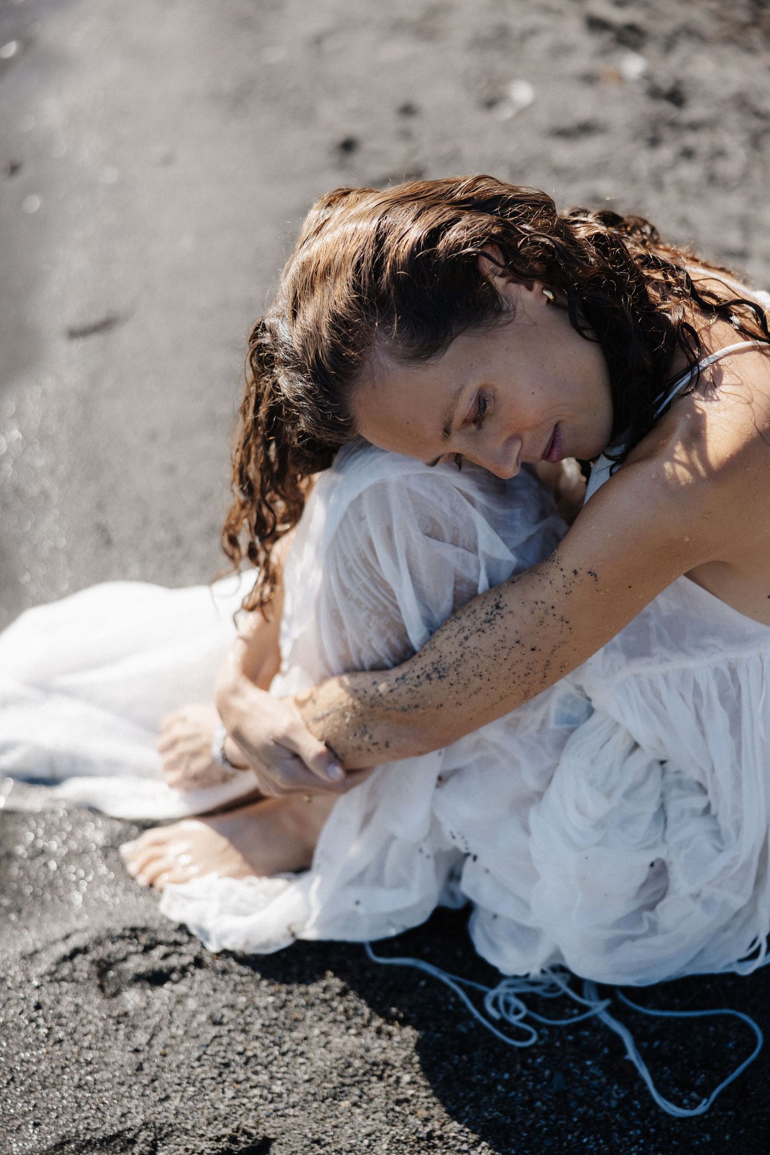 Woman in wet white dress sitting on dark sand beach, hugging knees with eyes closed.
