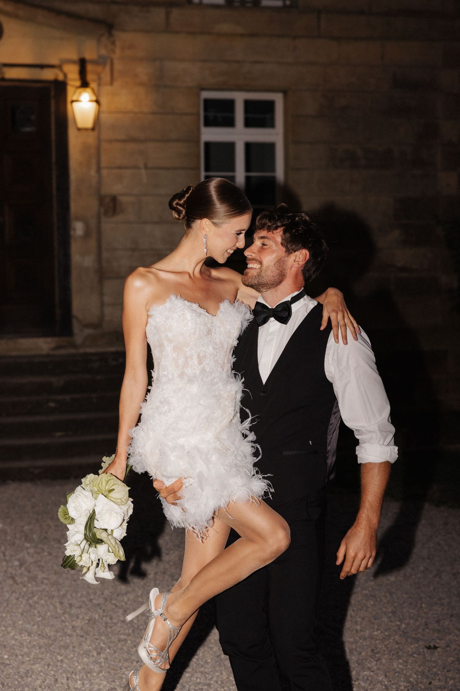 Couple embracing at night outside stone building, bride in white feather mini dress holding bouquet.