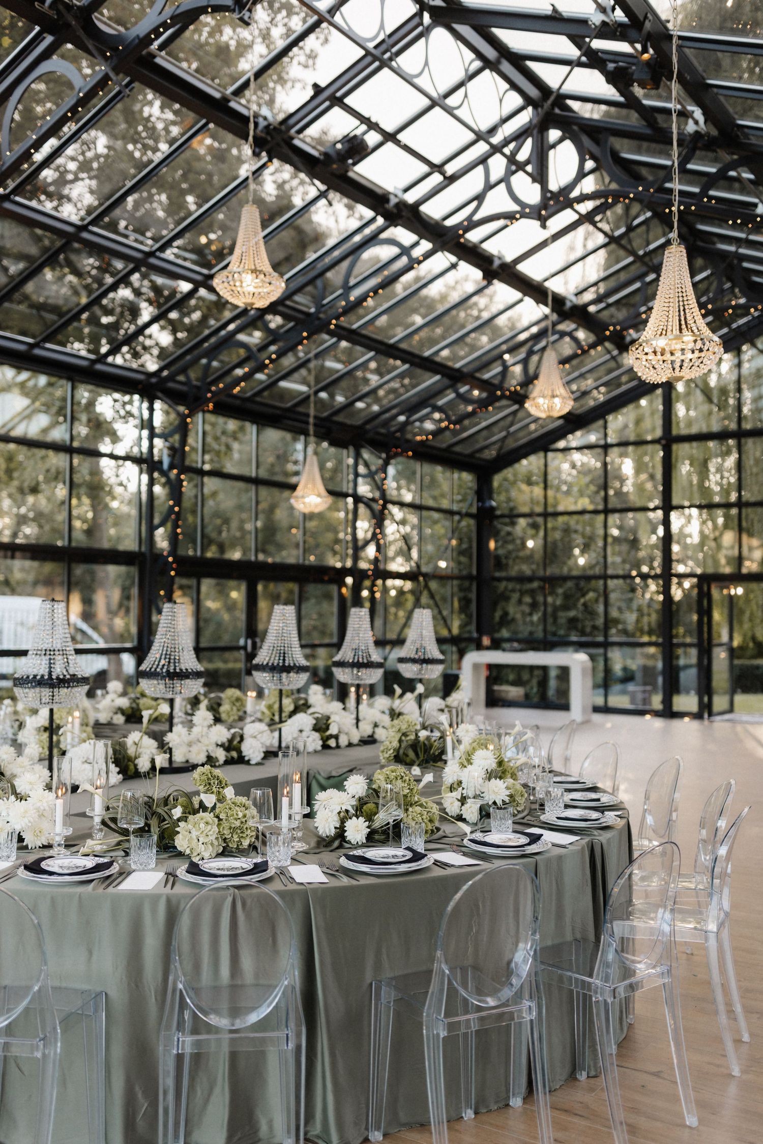 Elegant greenhouse wedding reception with crystal chandeliers, ghost chairs, and white floral centerpieces.
