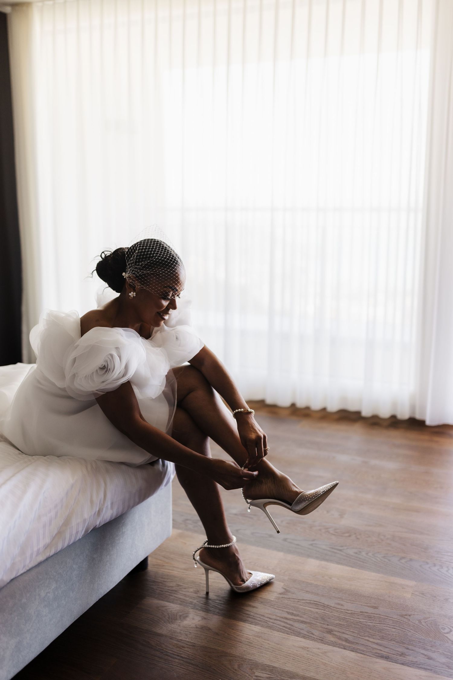 Black bride in white tulle wedding dress and birdcage veil fastens her strappy heels on bed.