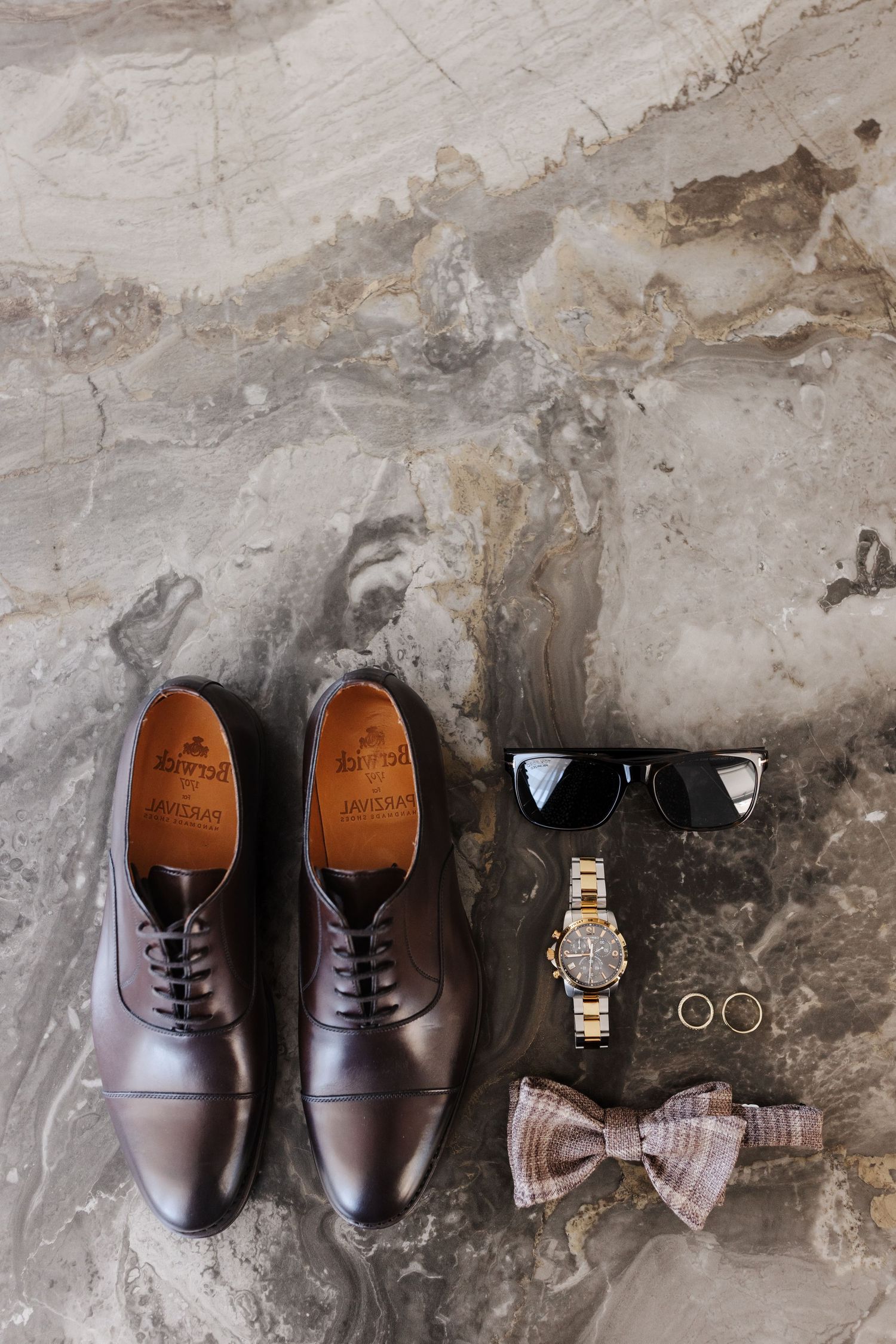 Groom essentials flat lay featuring brown Oxford shoes, sunglasses, gold watch, rings, and bow tie on marble.