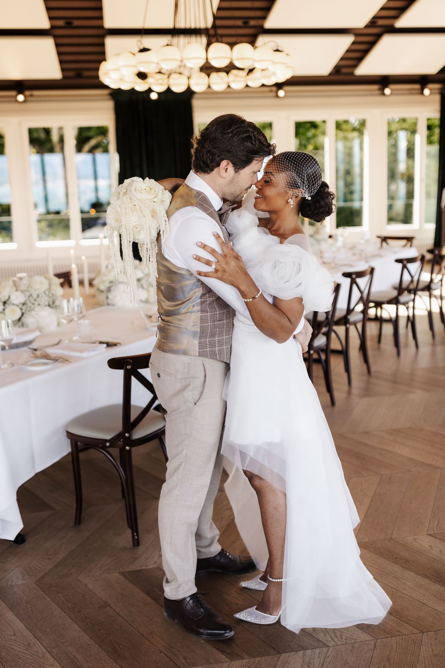 Interracial couple sharing romantic embrace in elegant wedding reception hall with chandelier.