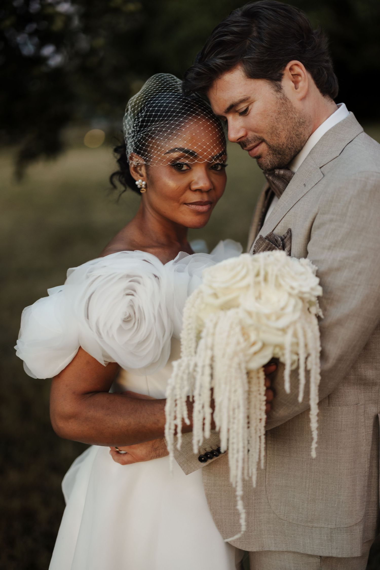 Bride in birdcage veil holding white hydrangea bouquet leaning close to groom in beige suit outdoors at dusk.