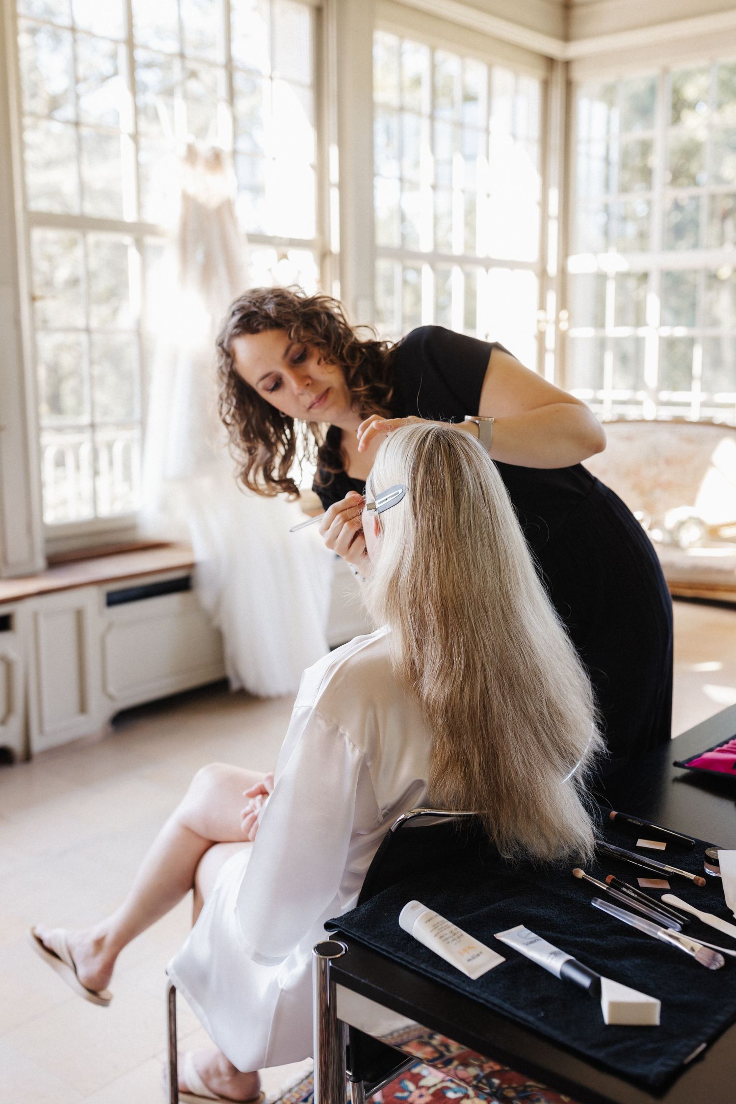 Hairstylist brushing blonde hair of bride in white robe by bright windows.