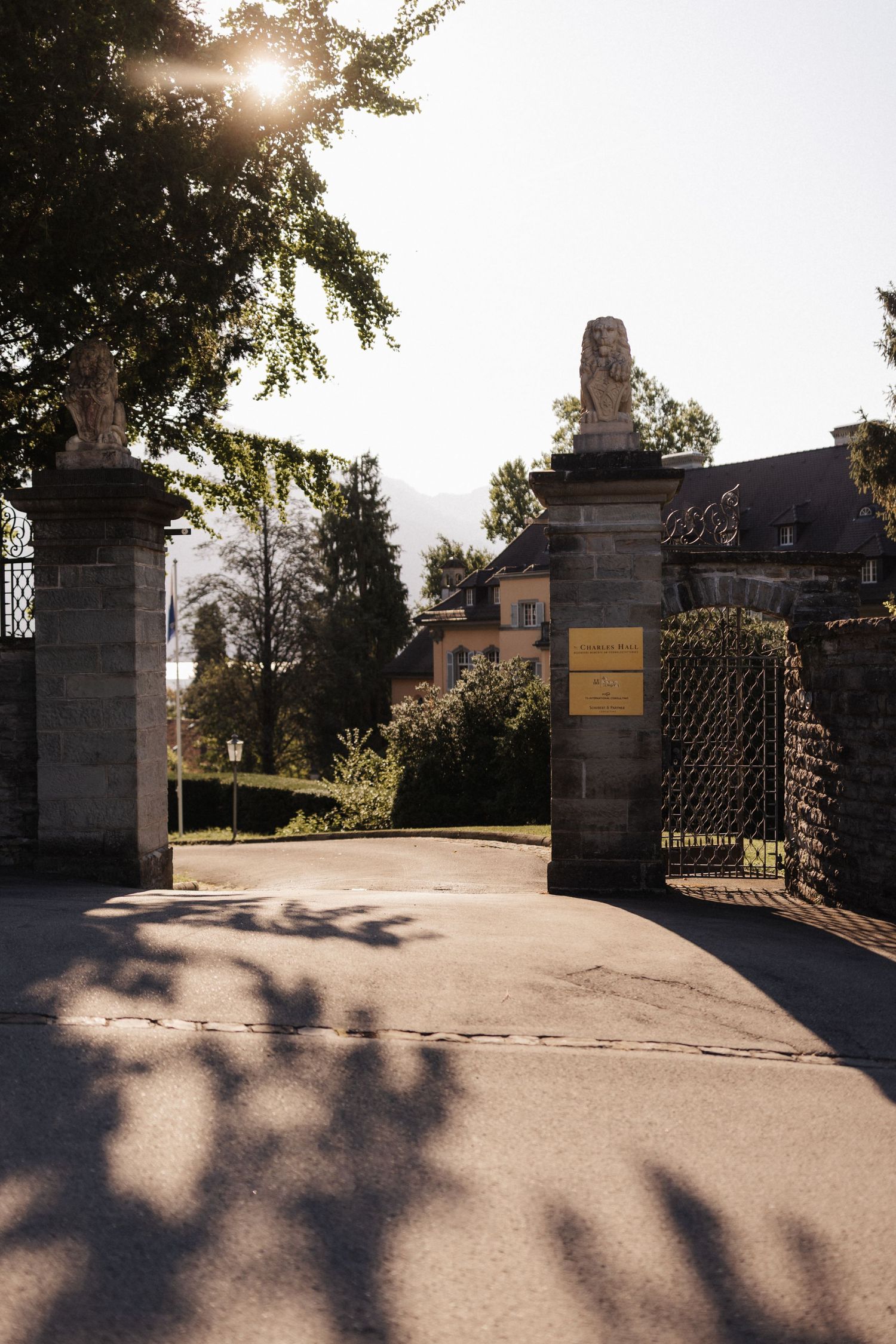 Sunlit ornate stone gate entrance with lion statues leading to an elegant historic estate.