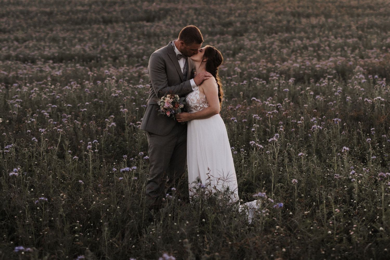 Newlyweds embrace romantically in a wildflower meadow during a warm and glowing sunset.