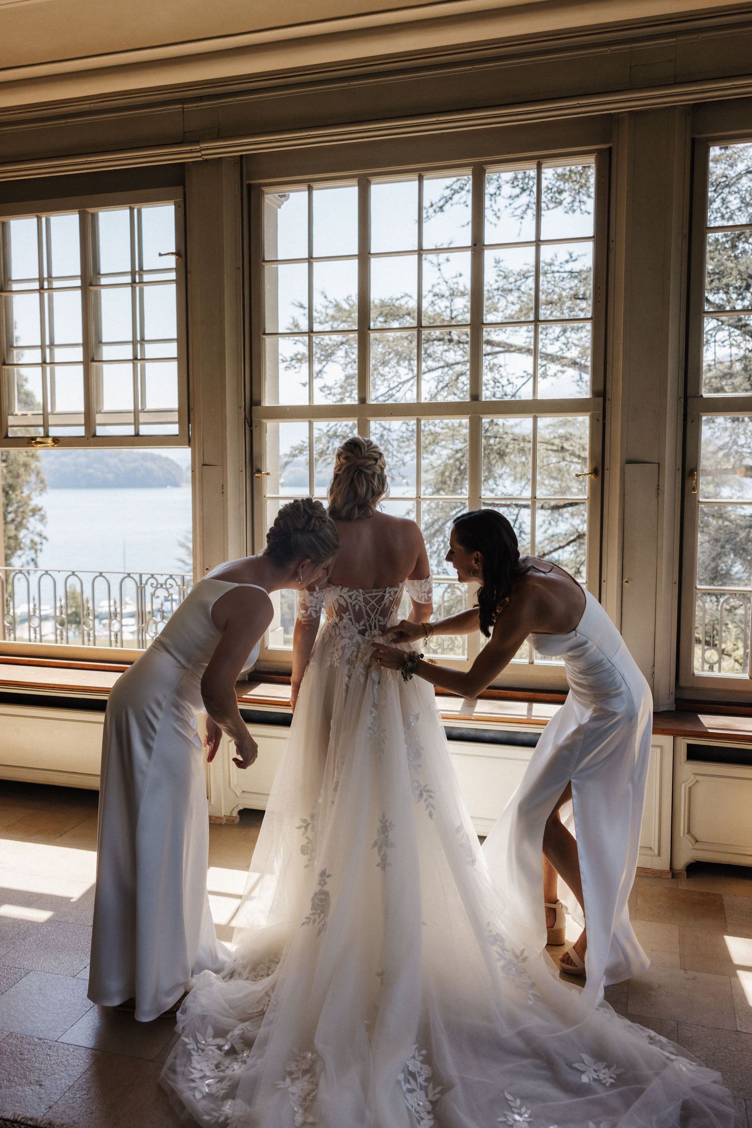 Bride in lace wedding gown assisted by two women in white dresses before large windows with lake view.