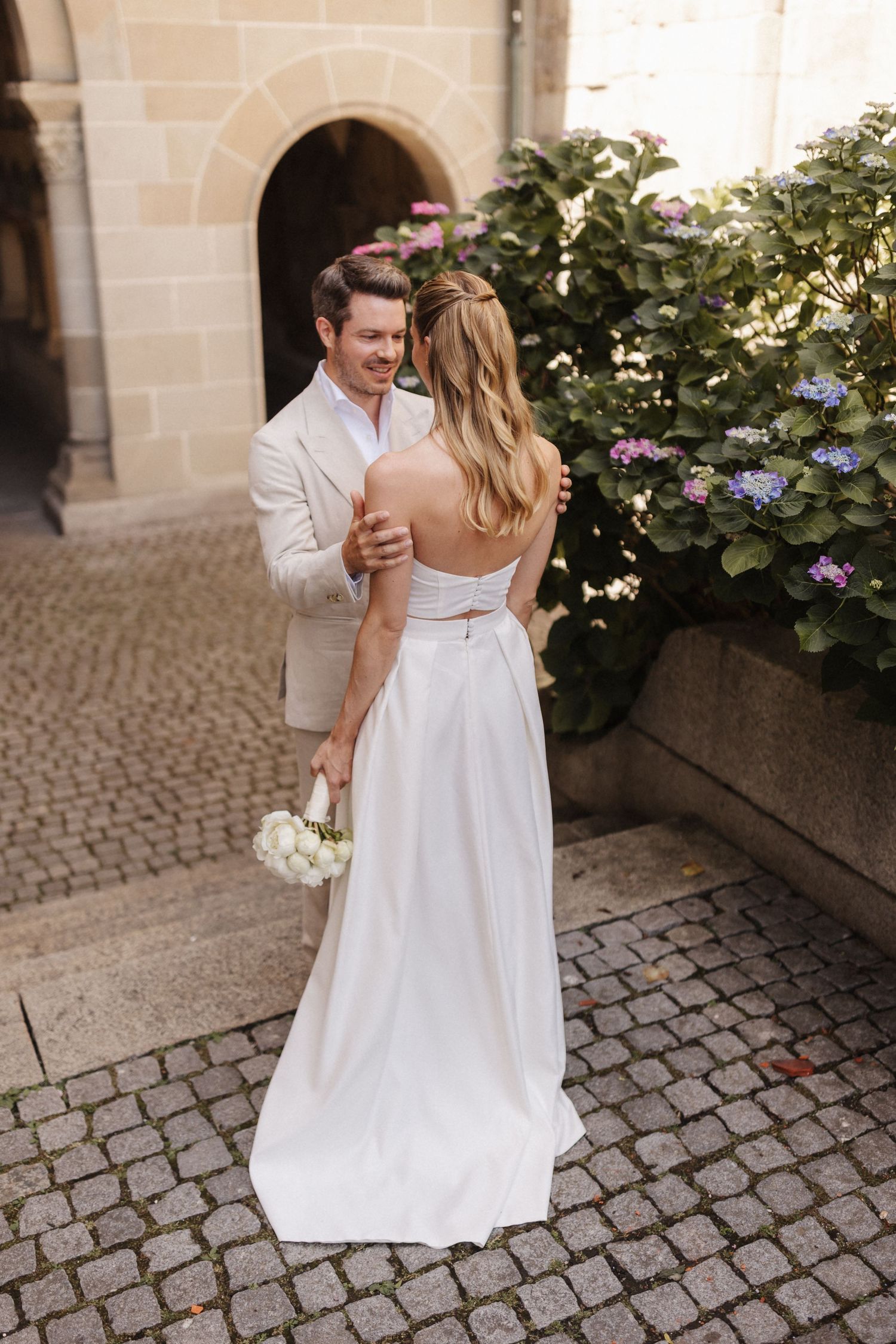 A groom in a beige suit embraces a bride in a white backless gown near blooming hydrangeas outdoors.