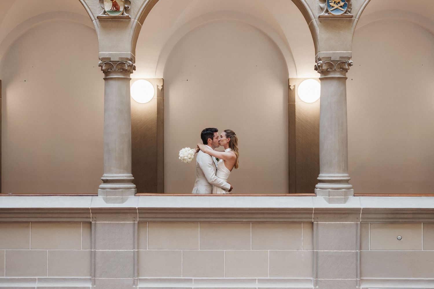 Bride and groom sharing romantic kiss under elegant arched colonnade in grand classical building on their wedding day.