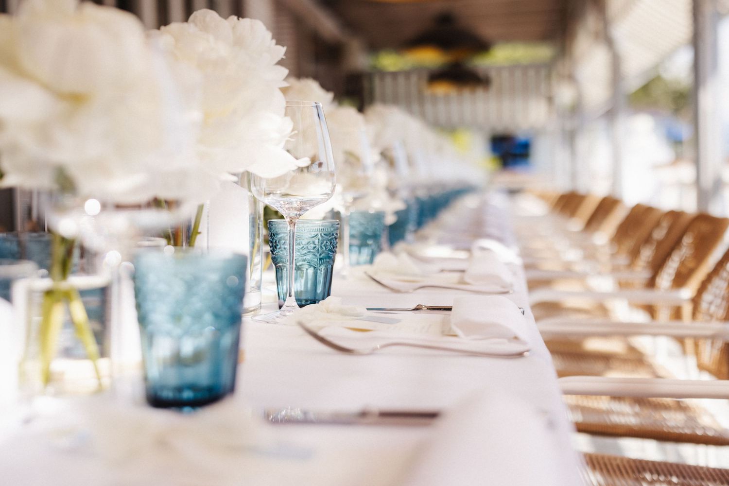 Close-up of wedding table setting featuring blue embossed glasses and white peony centerpieces on white linen.