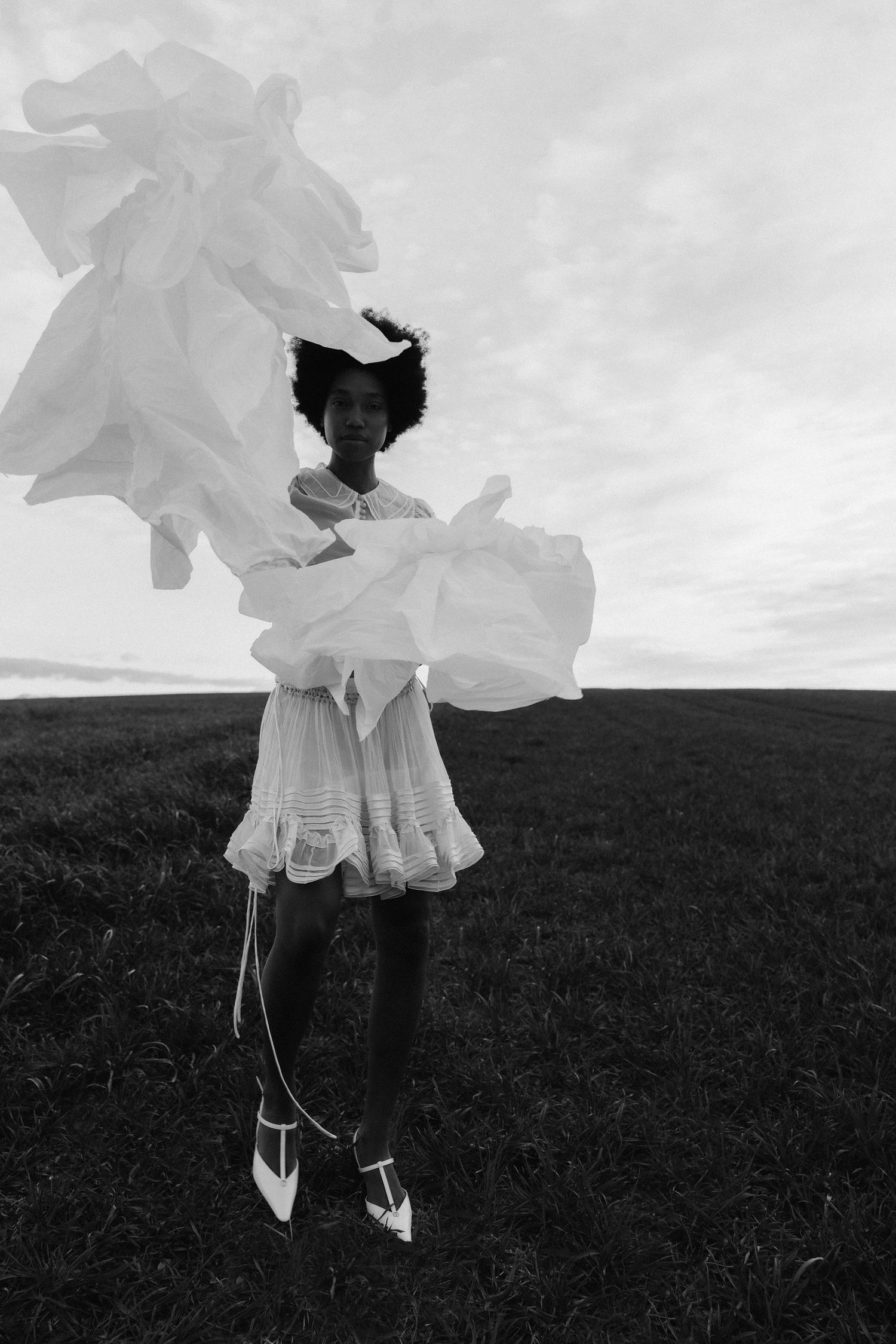 Black and white photo of a model in a white lace mini dress holding billowing white fabric in an open field.