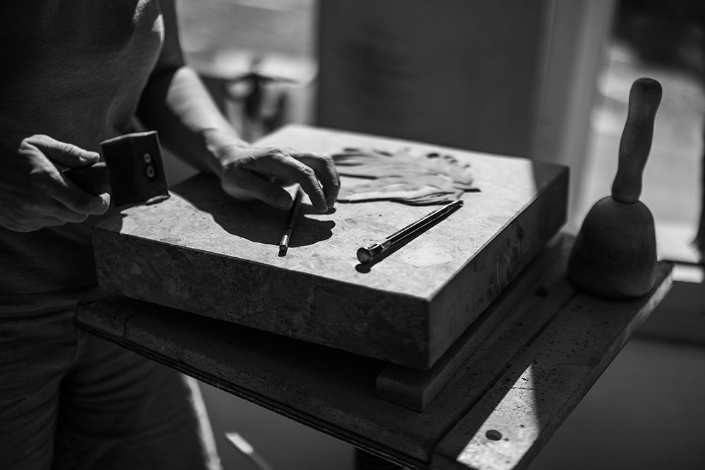 Close-up black and white image of sculptor's hands working on a stone slab with carving tools.