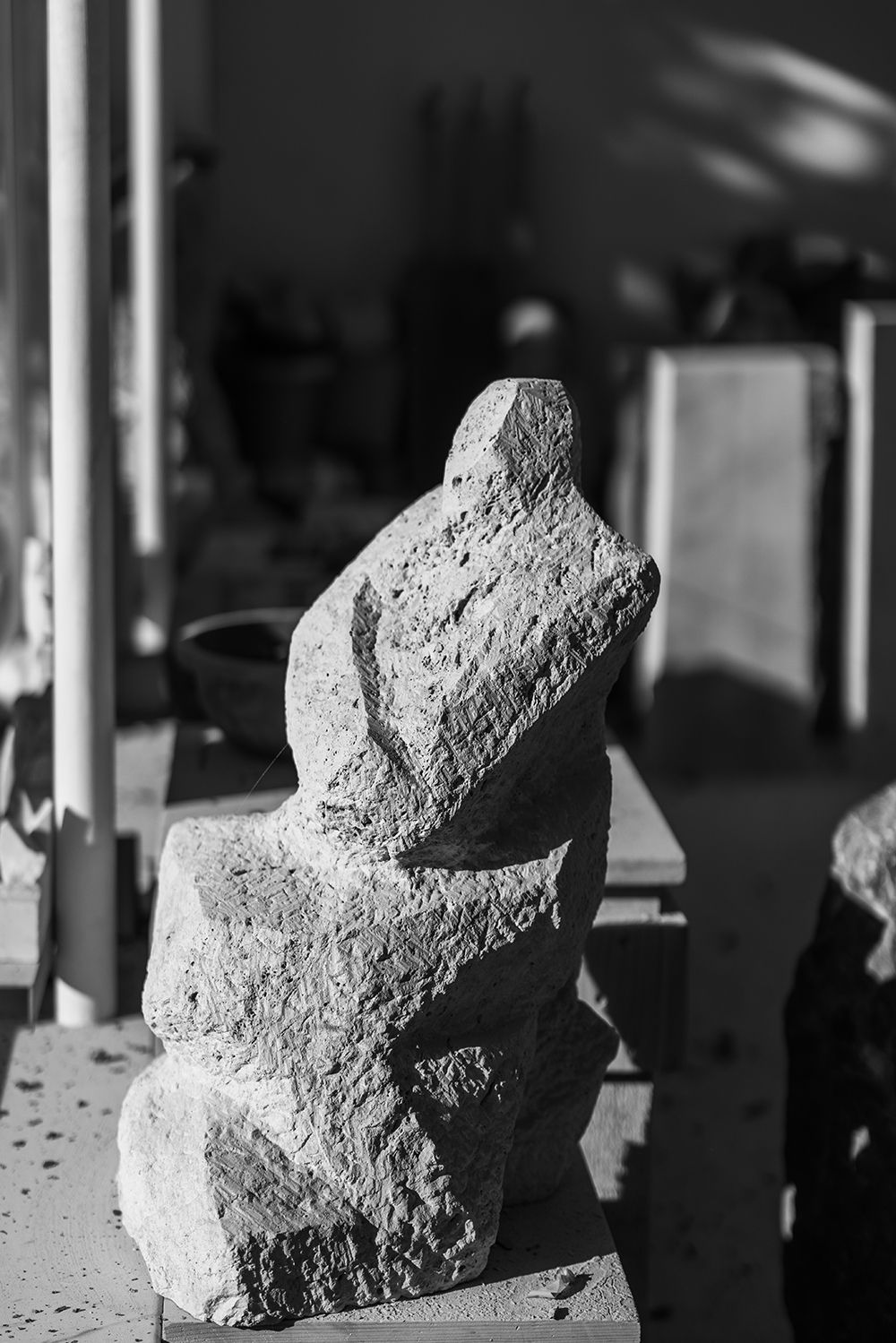 Black and white photo of a raw stone sculpture resembling a seated figure in a sculptor's workshop.