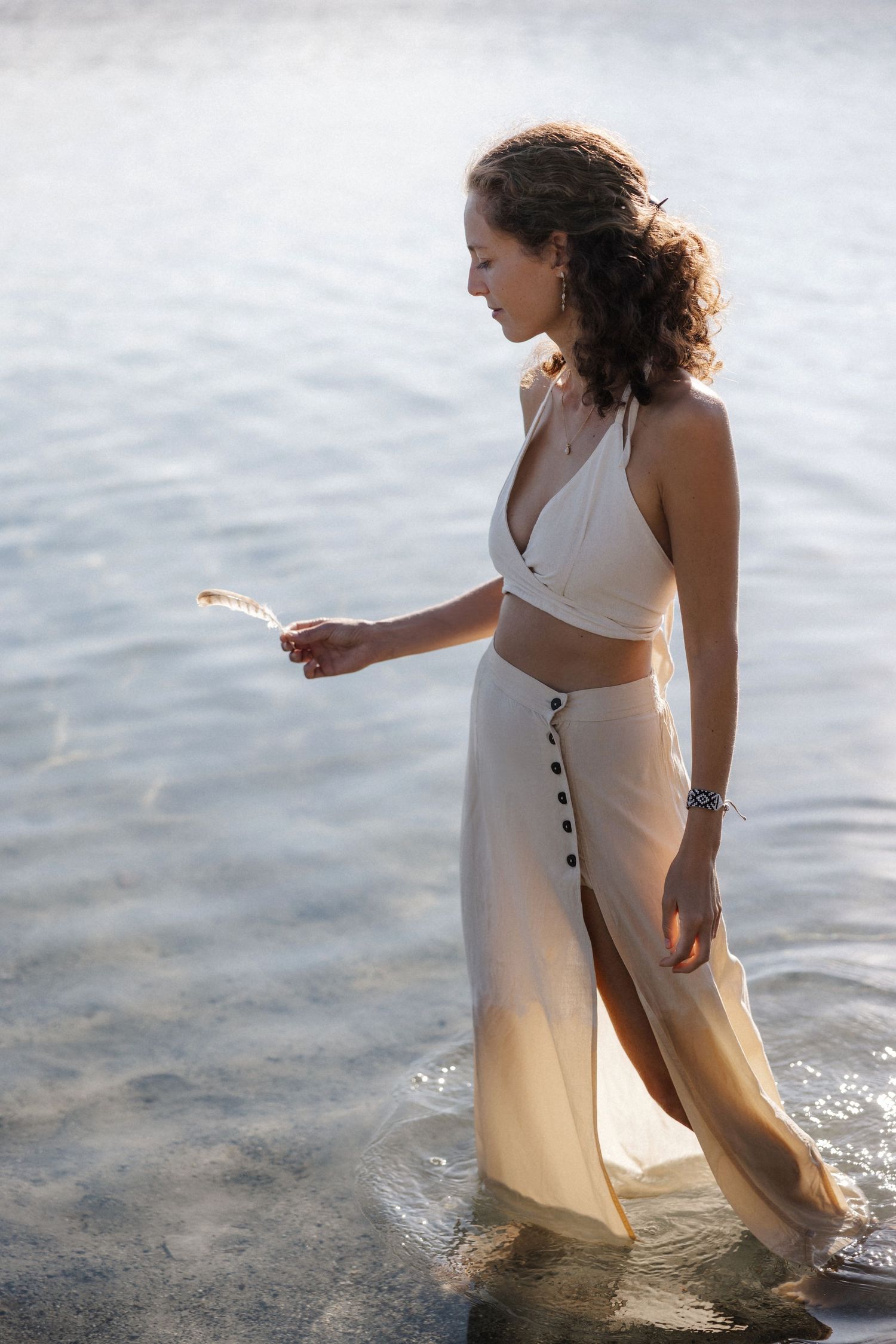 Woman in white crop top and cream skirt wading in shallow water, holding a small flower.