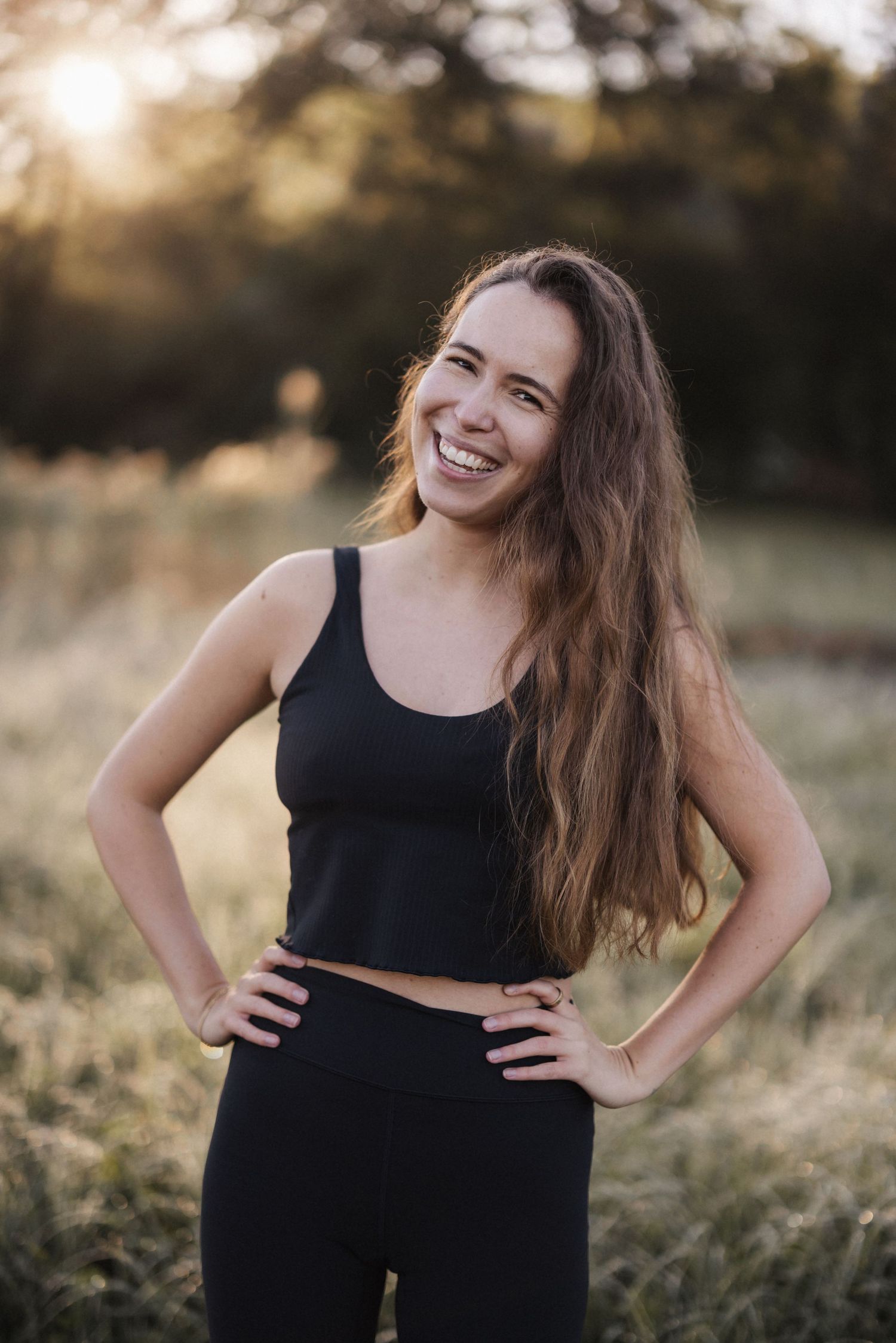 Smiling young woman with long hair in black crop top and leggings standing in a sunny outdoor field.