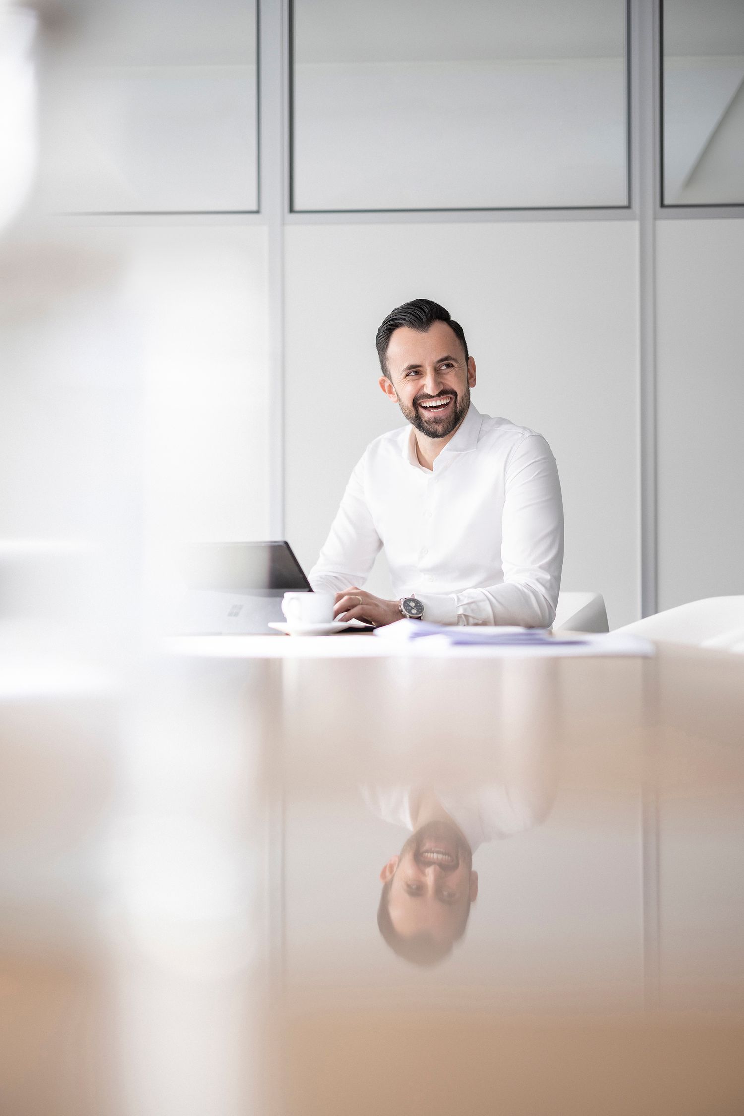 Happy bearded businessman in white shirt sitting at a reflective conference table with a laptop in a modern office.