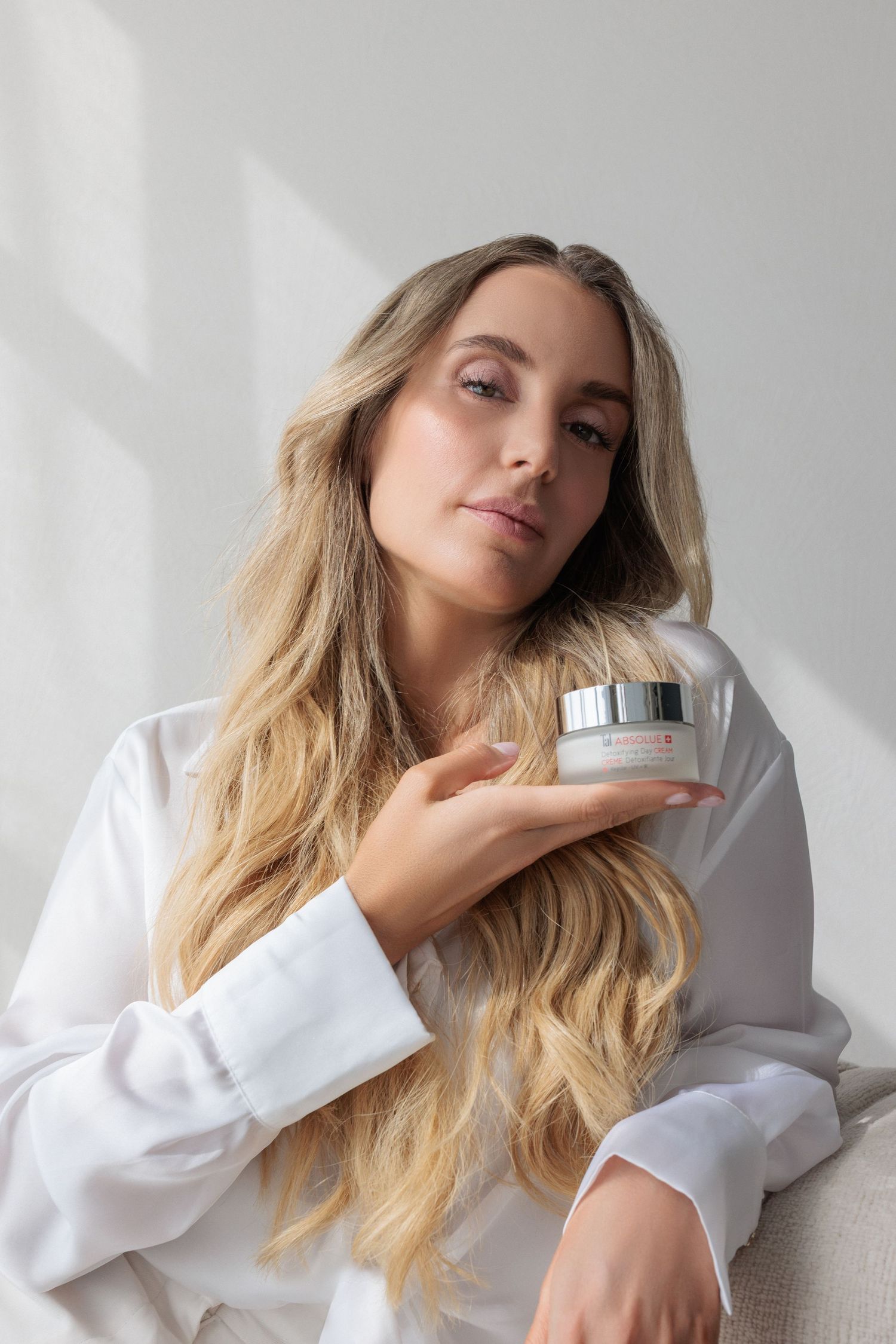 Blonde woman in white shirt holding skincare cream jar near face in bright minimalist setting.