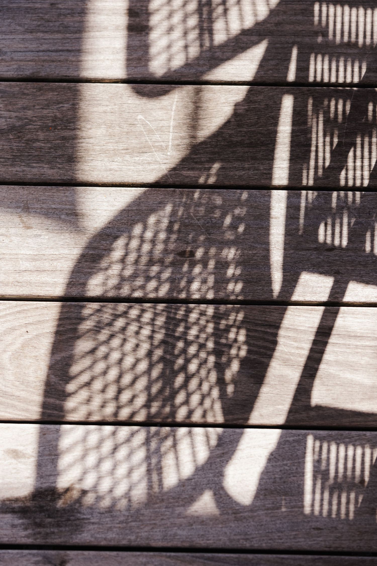 Artistic shadow of a wicker chair cast on weathered wooden deck planks in bright sunlight.