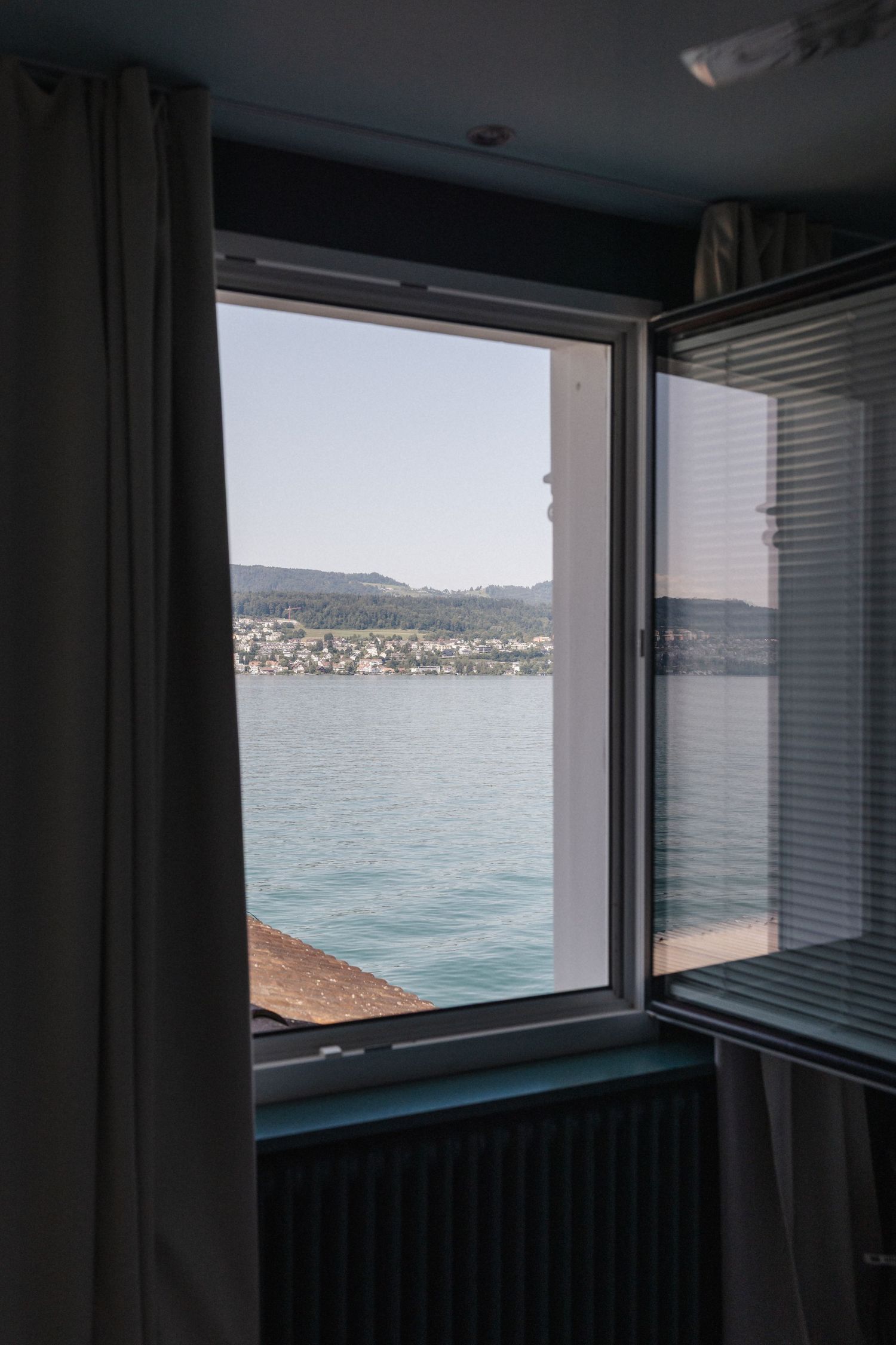 Open window with a serene lake and hillside town view from a hotel room.