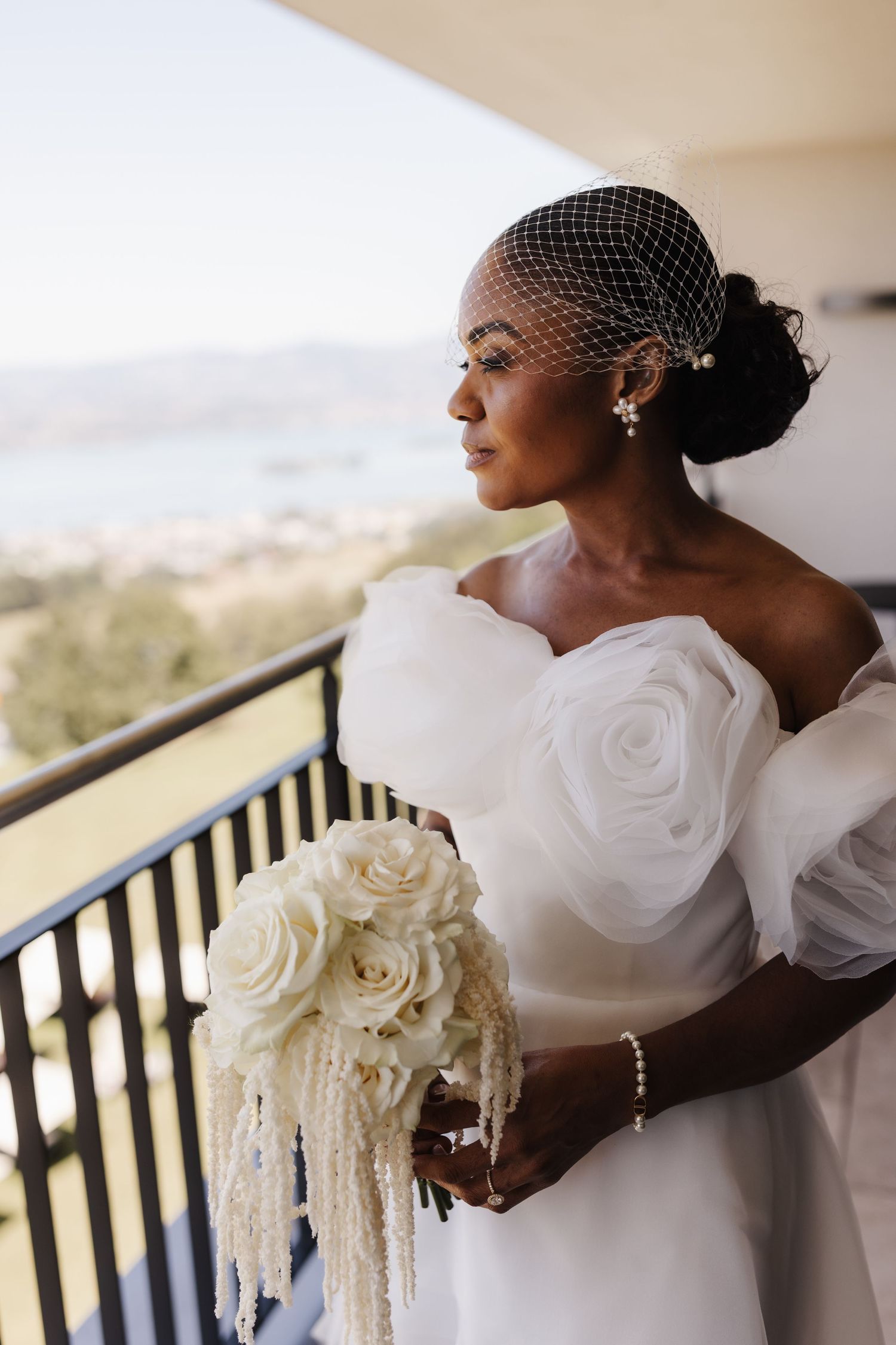 Black bride in white off-shoulder gown and birdcage veil holds white rose bouquet on balcony.