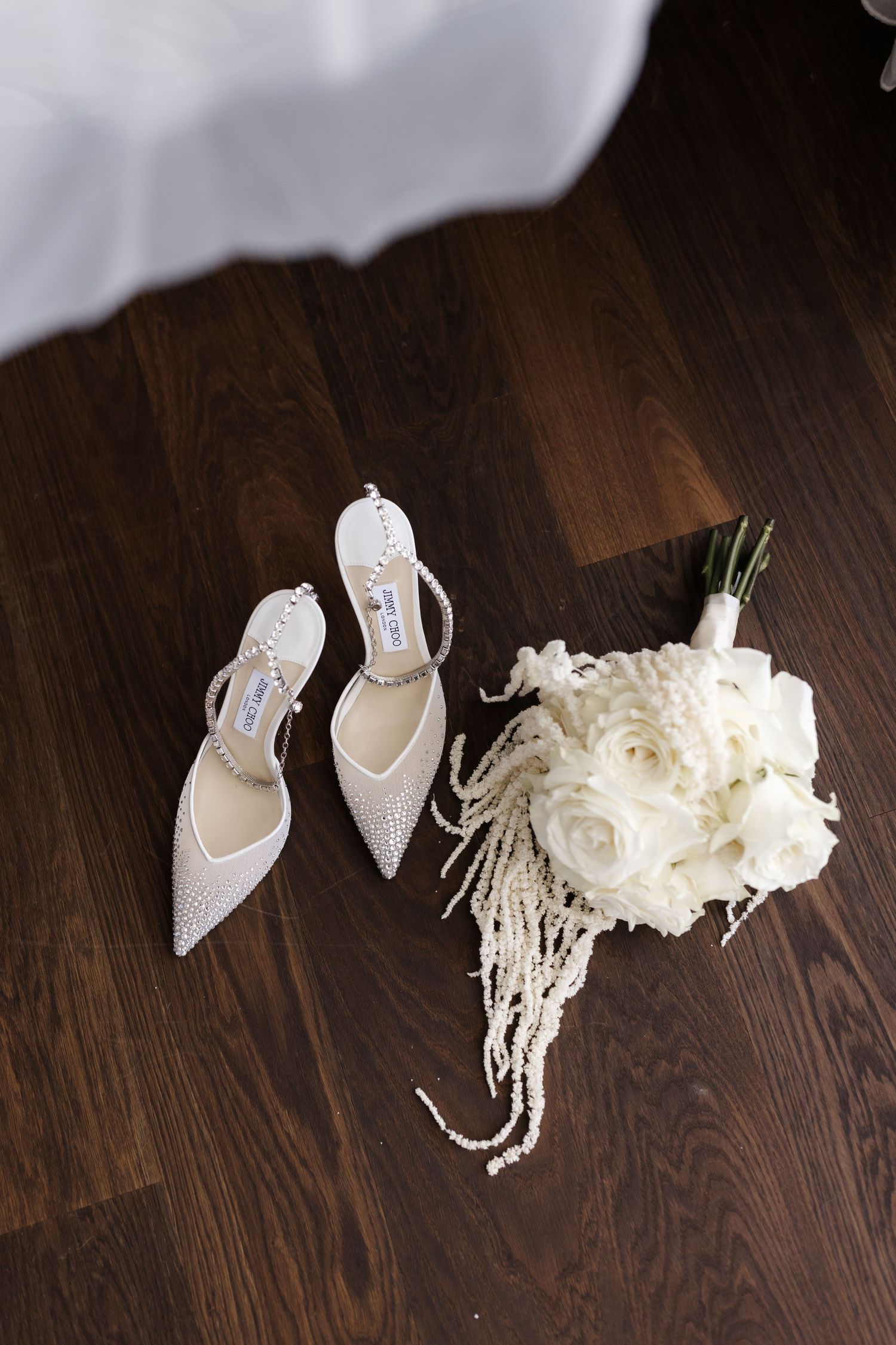 White crystal Jimmy Choo bridal heels beside a white rose bouquet with pearl details on wood floor.