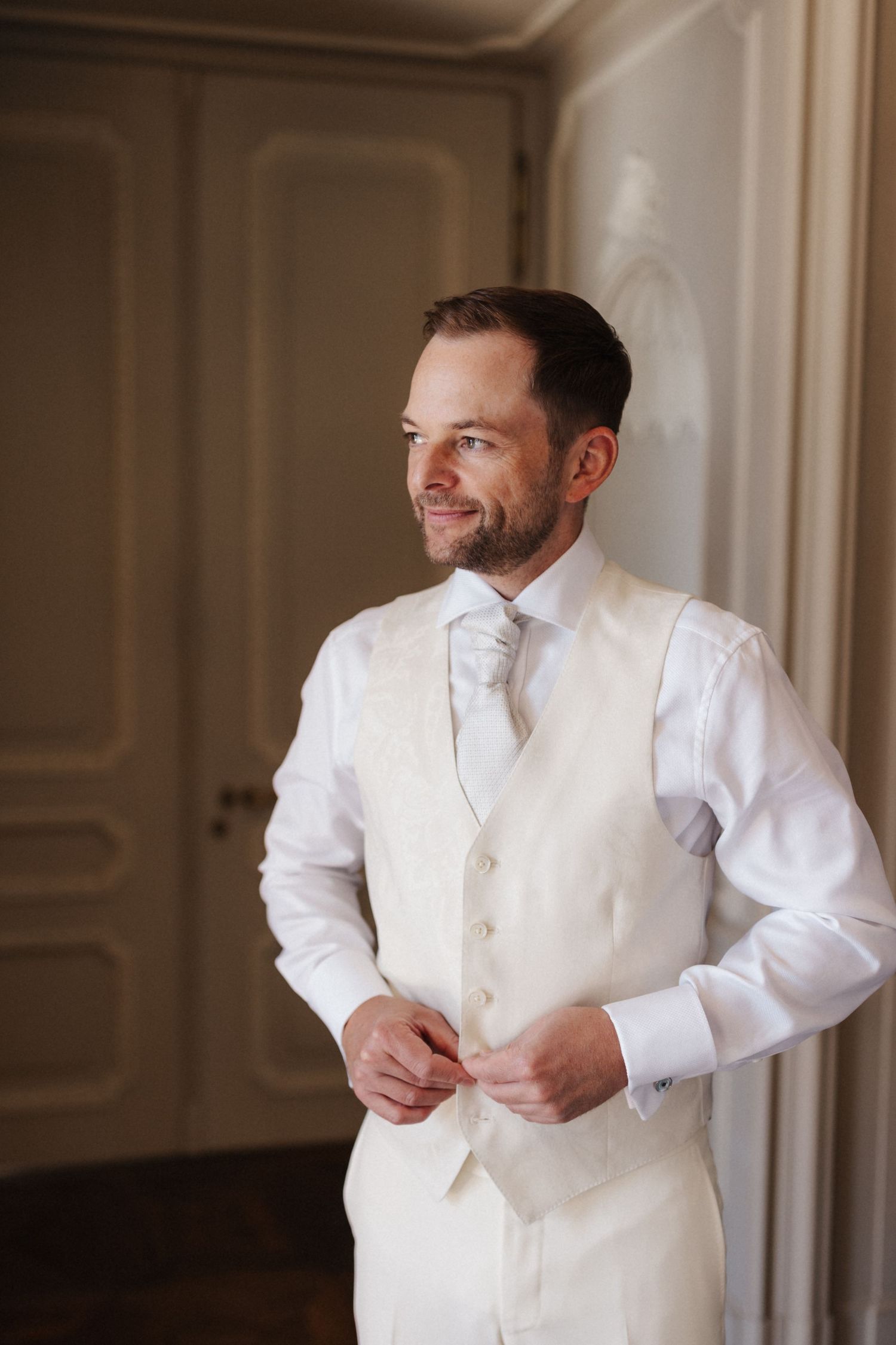 Smiling groom in white vest and cravat buttoning jacket in elegant room by window.
