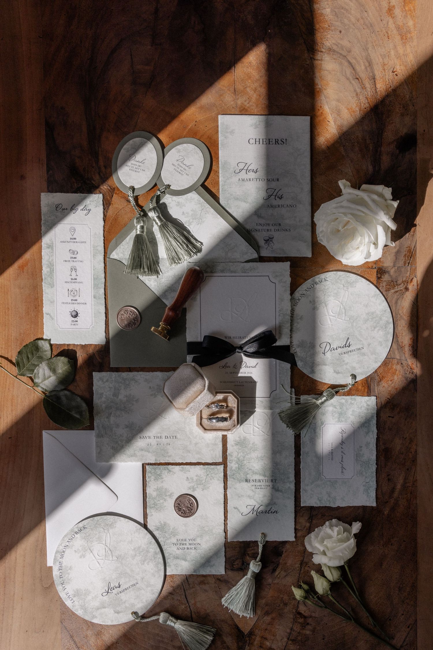 Overhead flatlay of a complete wedding stationery suite with tassels, wax seals, and white roses on wood.