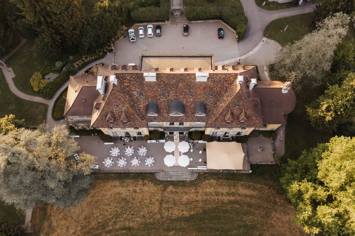 Aerial view of a historic yellow villa with terracotta roof surrounded by gardens and outdoor dining tables.