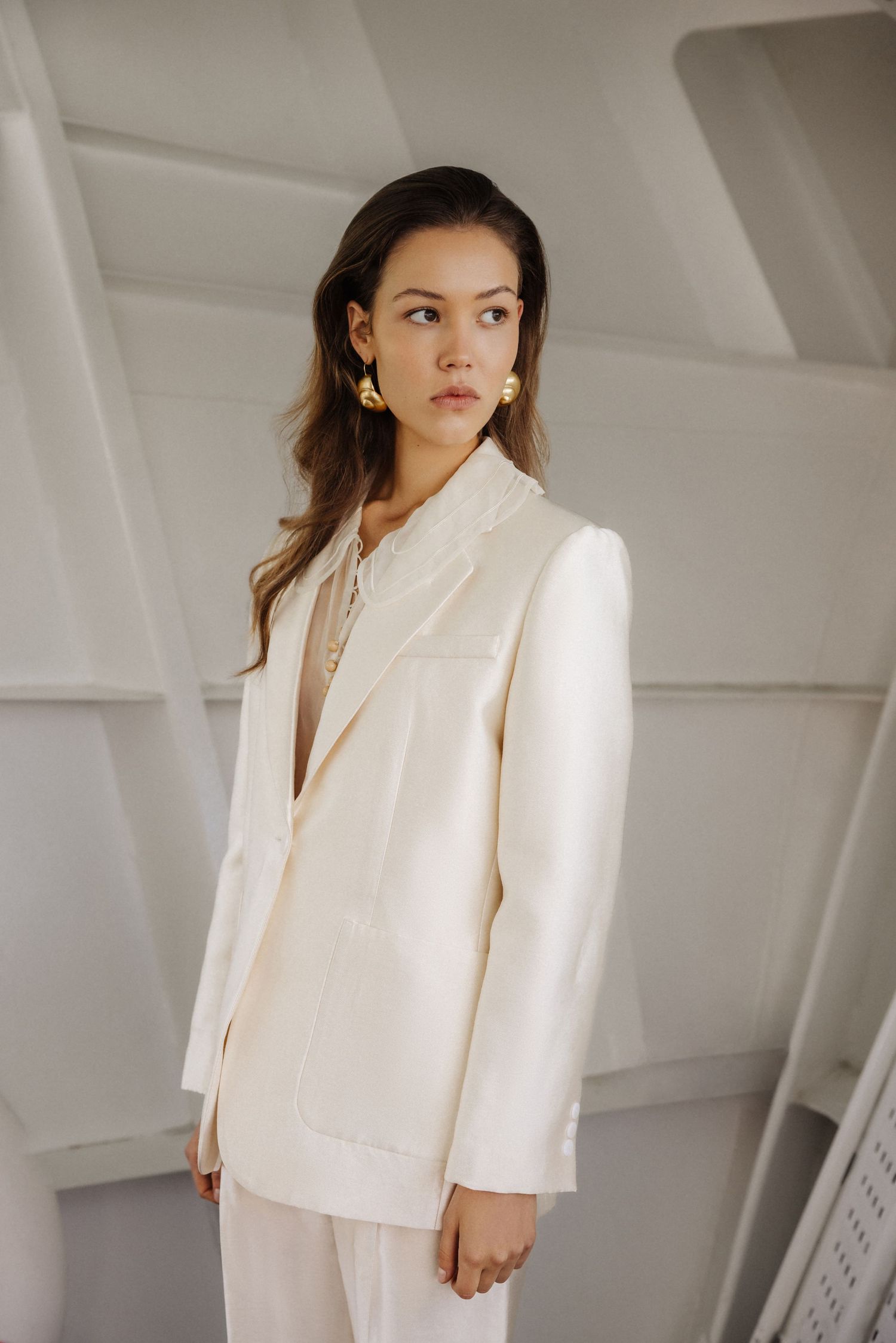 Woman in elegant white blazer suit with gold earrings posing in modern white interior.