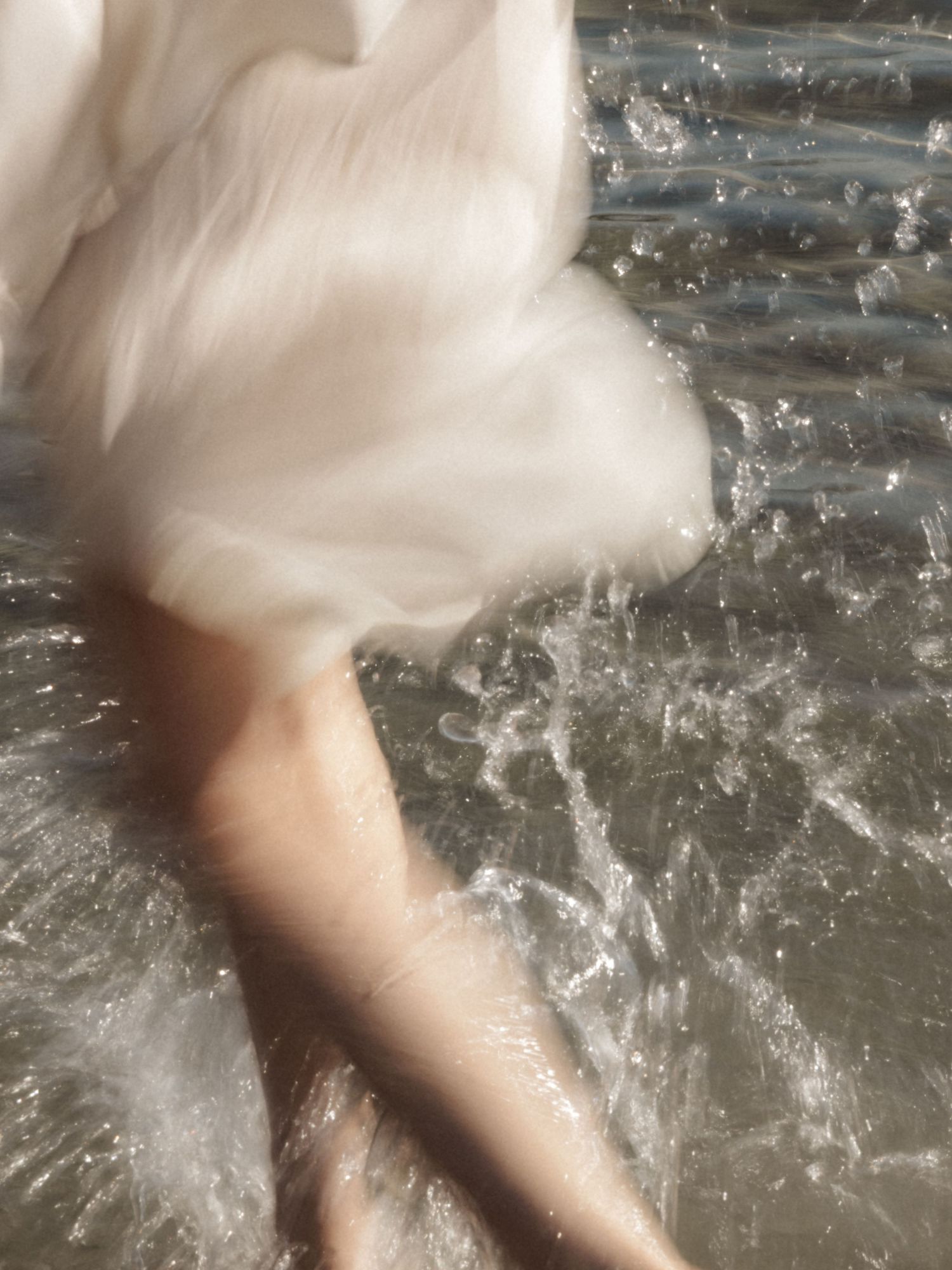 Woman in white dress with legs in splashing clear shallow water at beach.