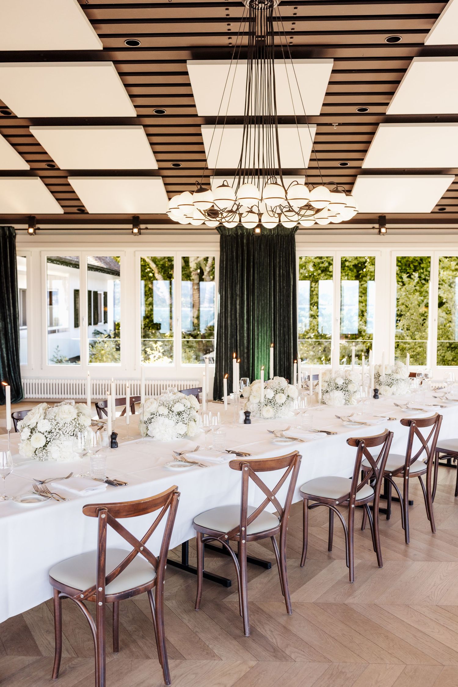 Bright wedding reception room featuring long white-draped table, wooden chairs, coffered ceiling, chandelier, and garden views.