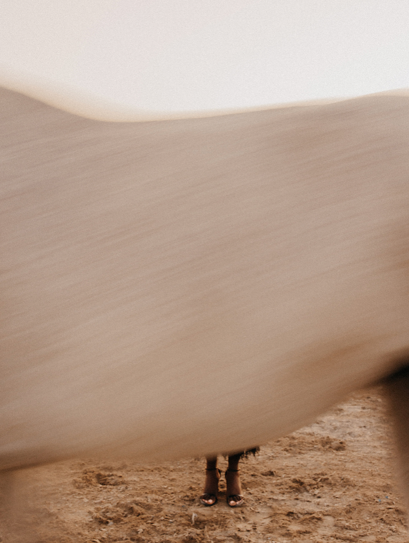 Minimalist fashion photo of person obscured by flowing beige fabric billowing in desert wind.