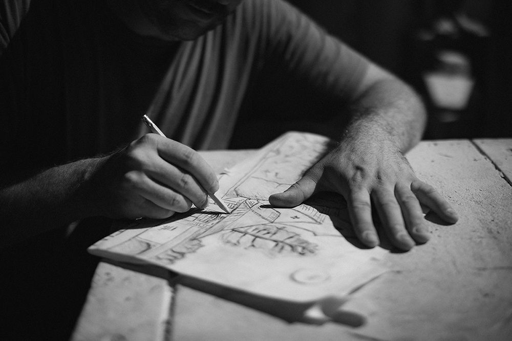 Black and white photo of hands sketching detailed artwork on paper at a rustic table.