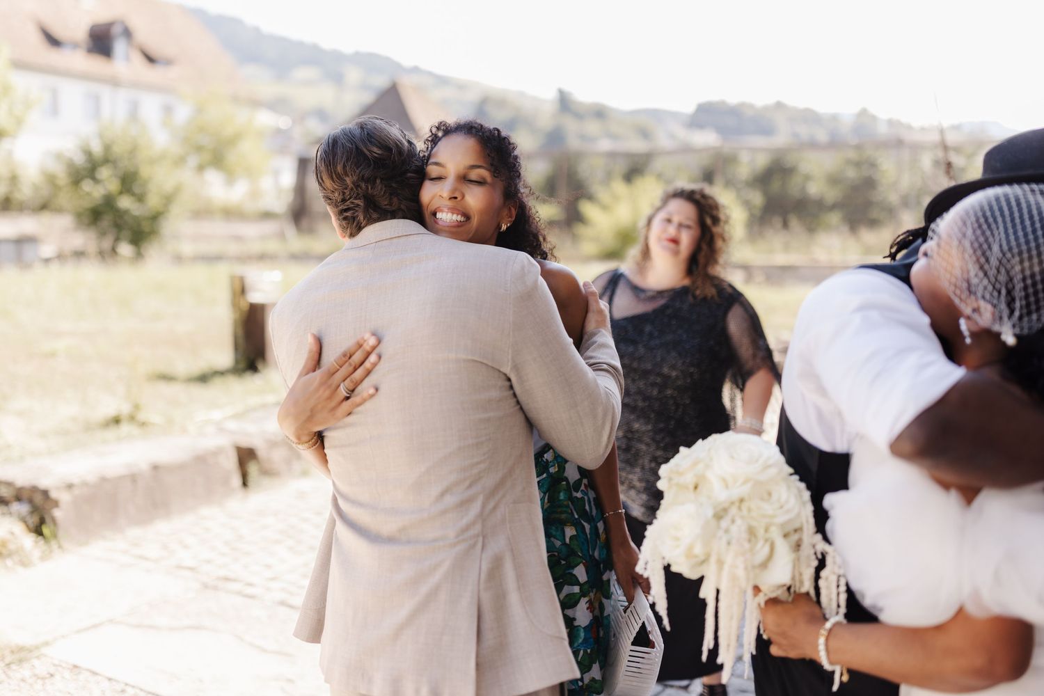 Bride in white wedding dress hugs guest outdoors while another woman laughs in background at wedding.