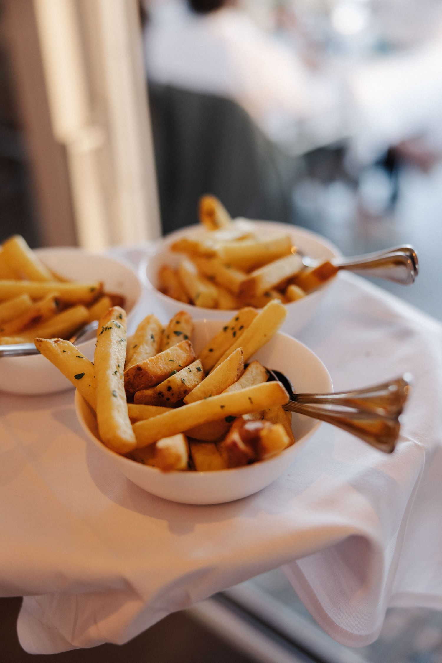Three white bowls of seasoned french fries served with silver spoons on a white cloth.