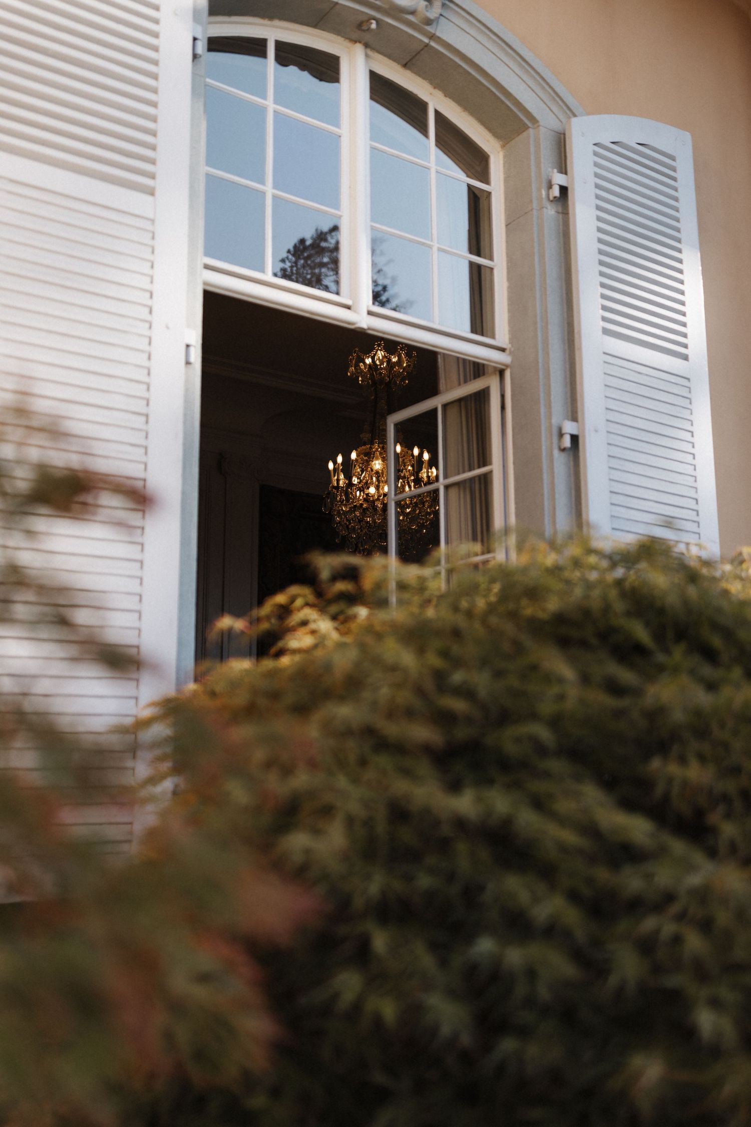 Elegant arched window with white shutters revealing a chandelier inside a classic mansion.