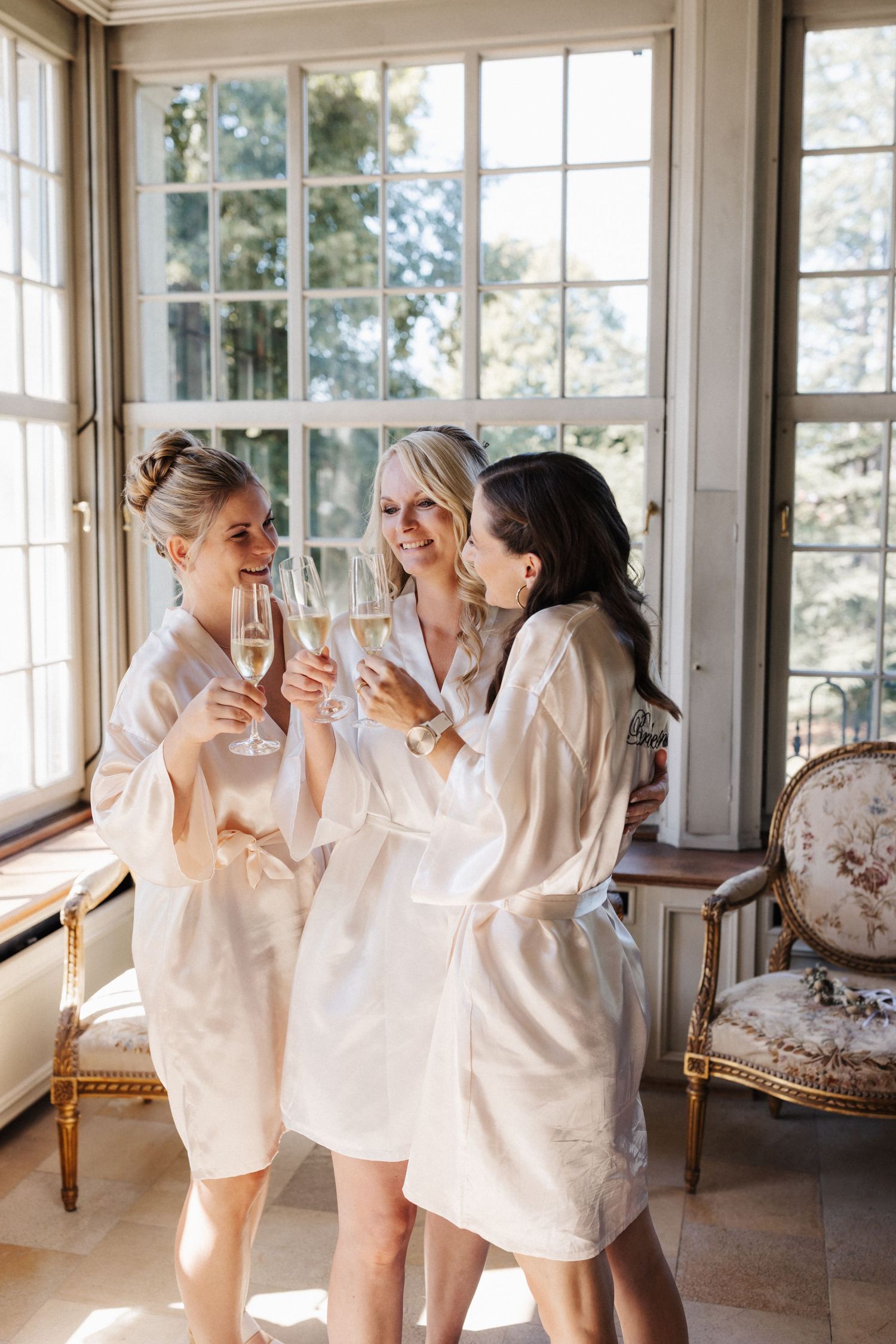 Three women in white satin robes clinking champagne glasses near large windows in an elegant room.