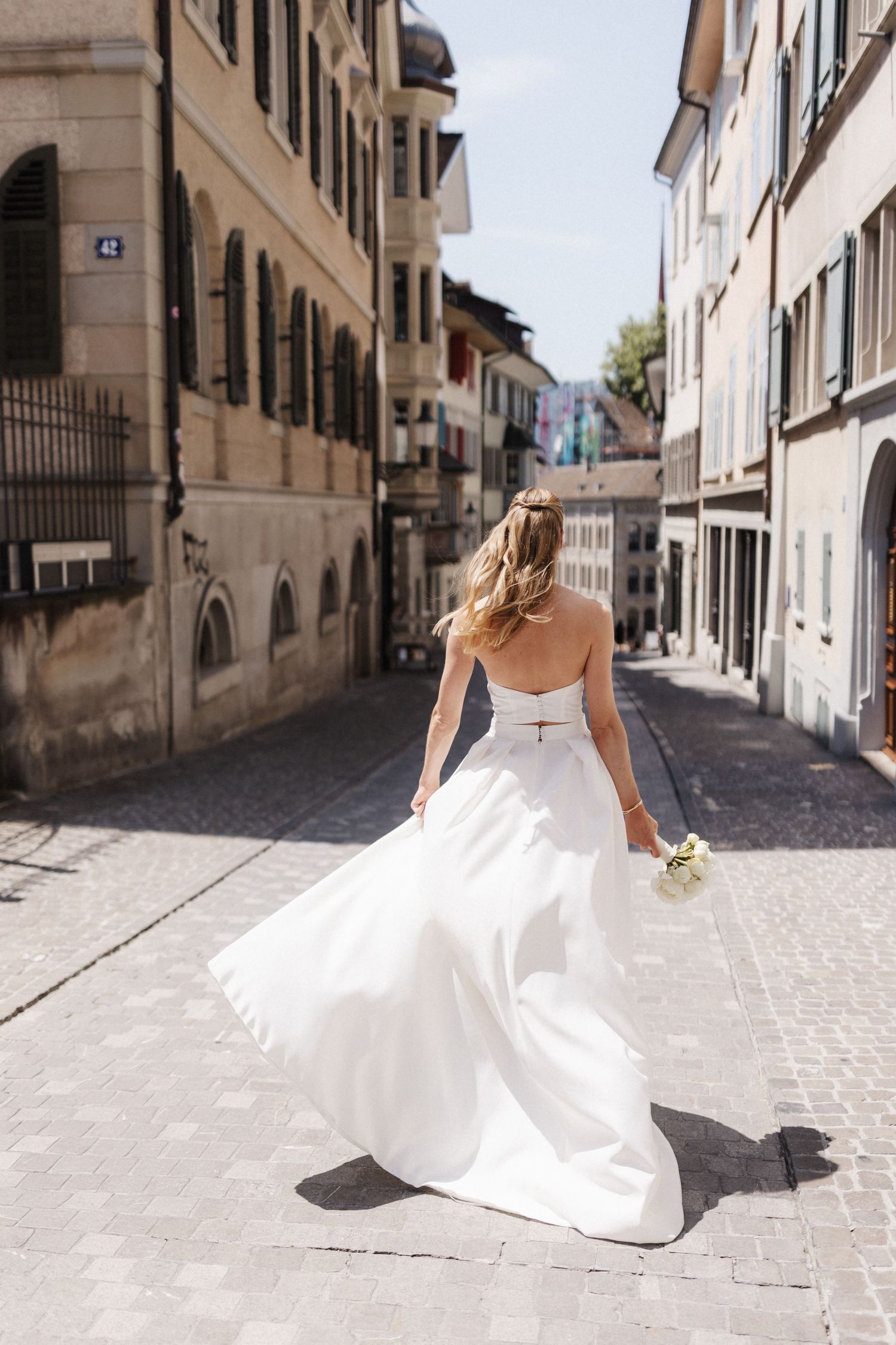 A bride in a flowing white two-piece gown walks away down a sunny cobblestone European street.