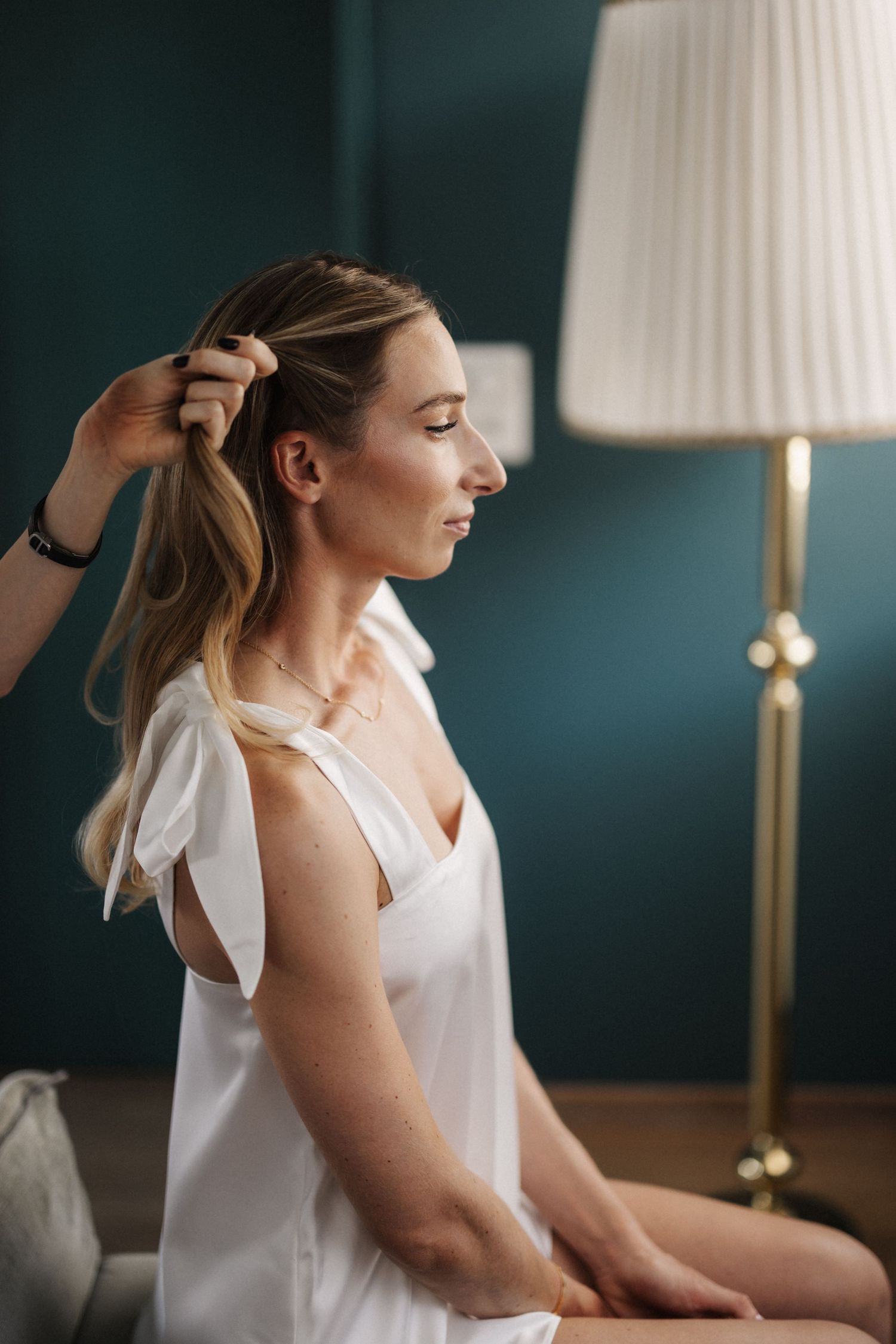 Hairstylist pinning blonde bride's hair as she sits in white dress.