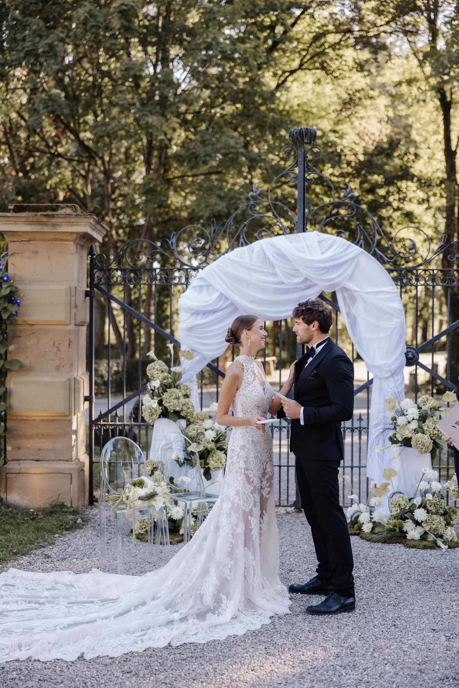 Bride in lace gown and groom in tuxedo exchange vows before a white draped arch with white flowers.
