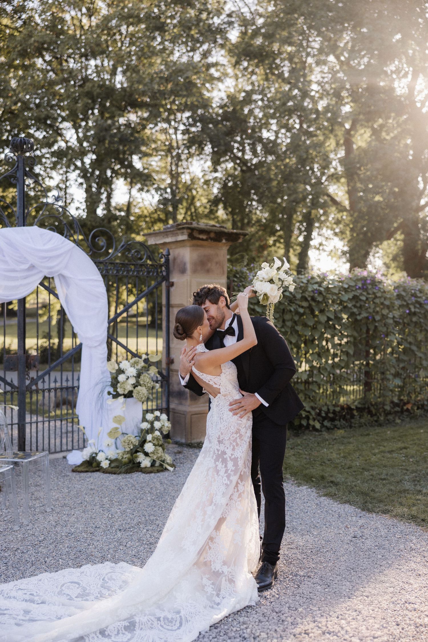 Bride and groom share a romantic kiss near ornate garden gates at golden hour sunset.