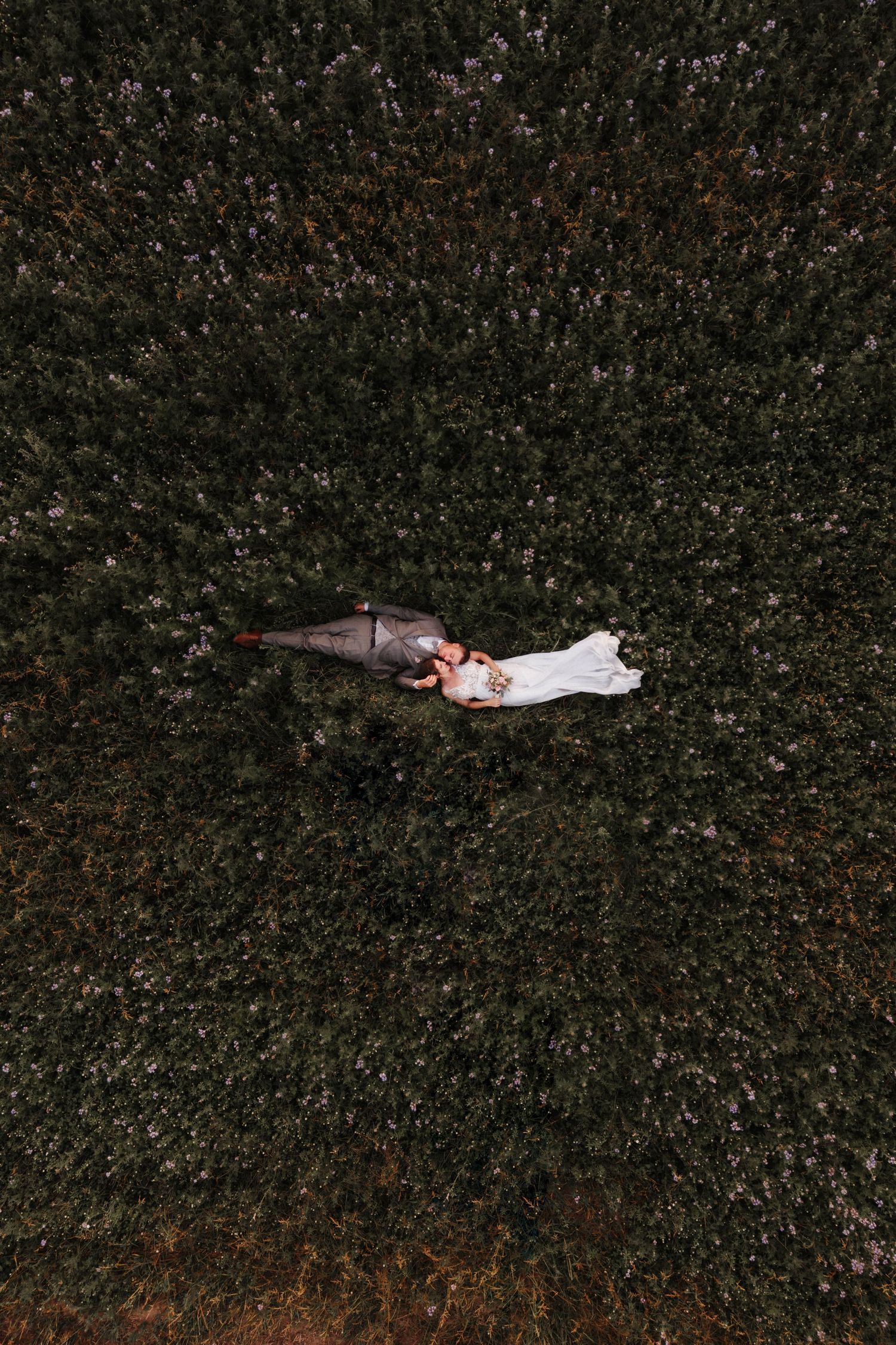 Aerial drone view of bride and groom lying together in a lush green wildflower field on their wedding day.