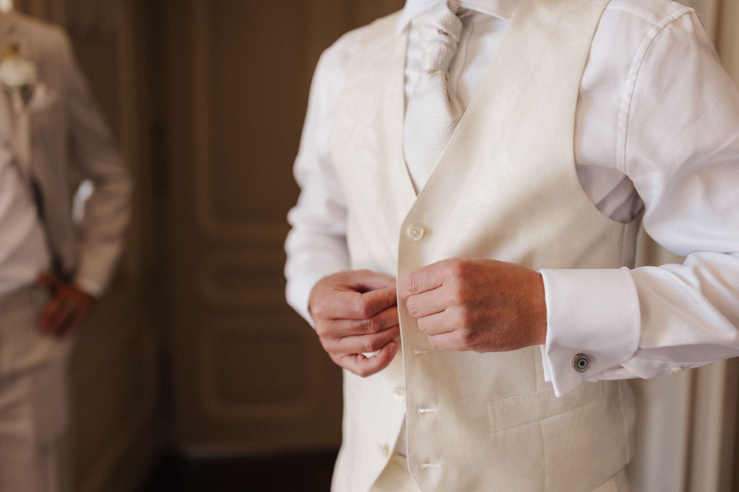 Groom in white dress shirt and ivory vest buttoning up while getting ready for wedding.
