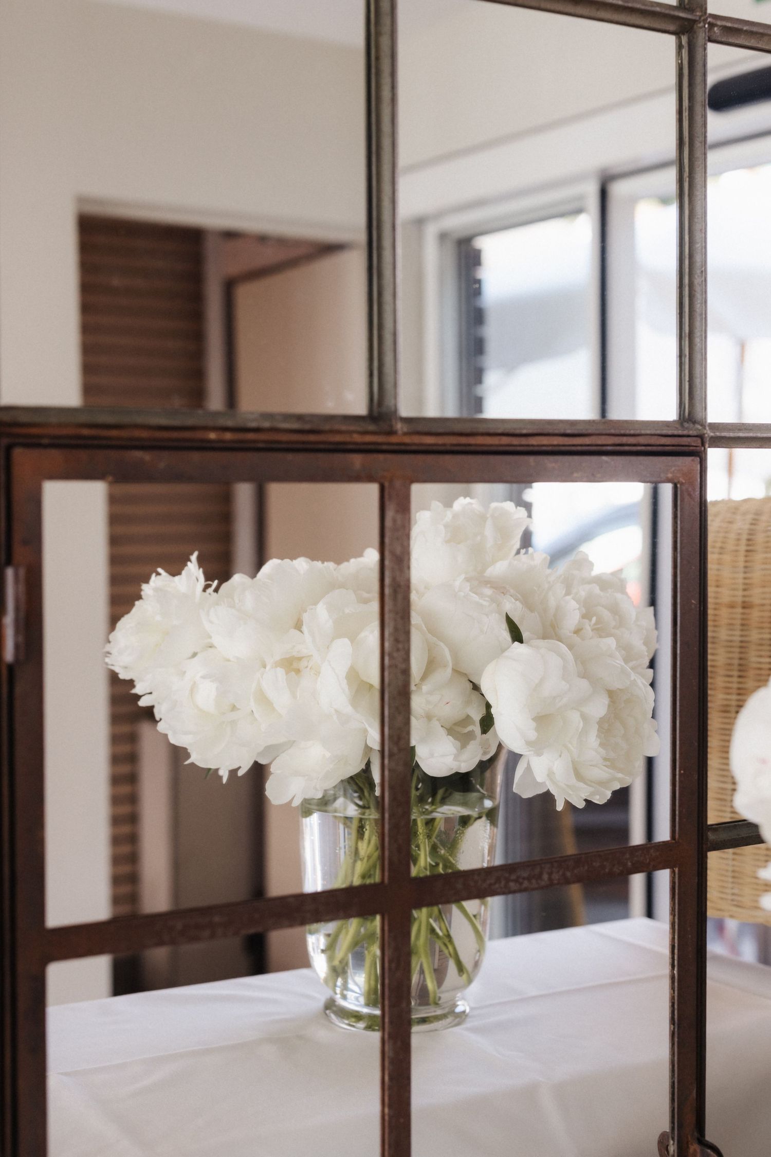 White peonies in glass vase displayed on white tablecloth beside decorative dark mirror frame.