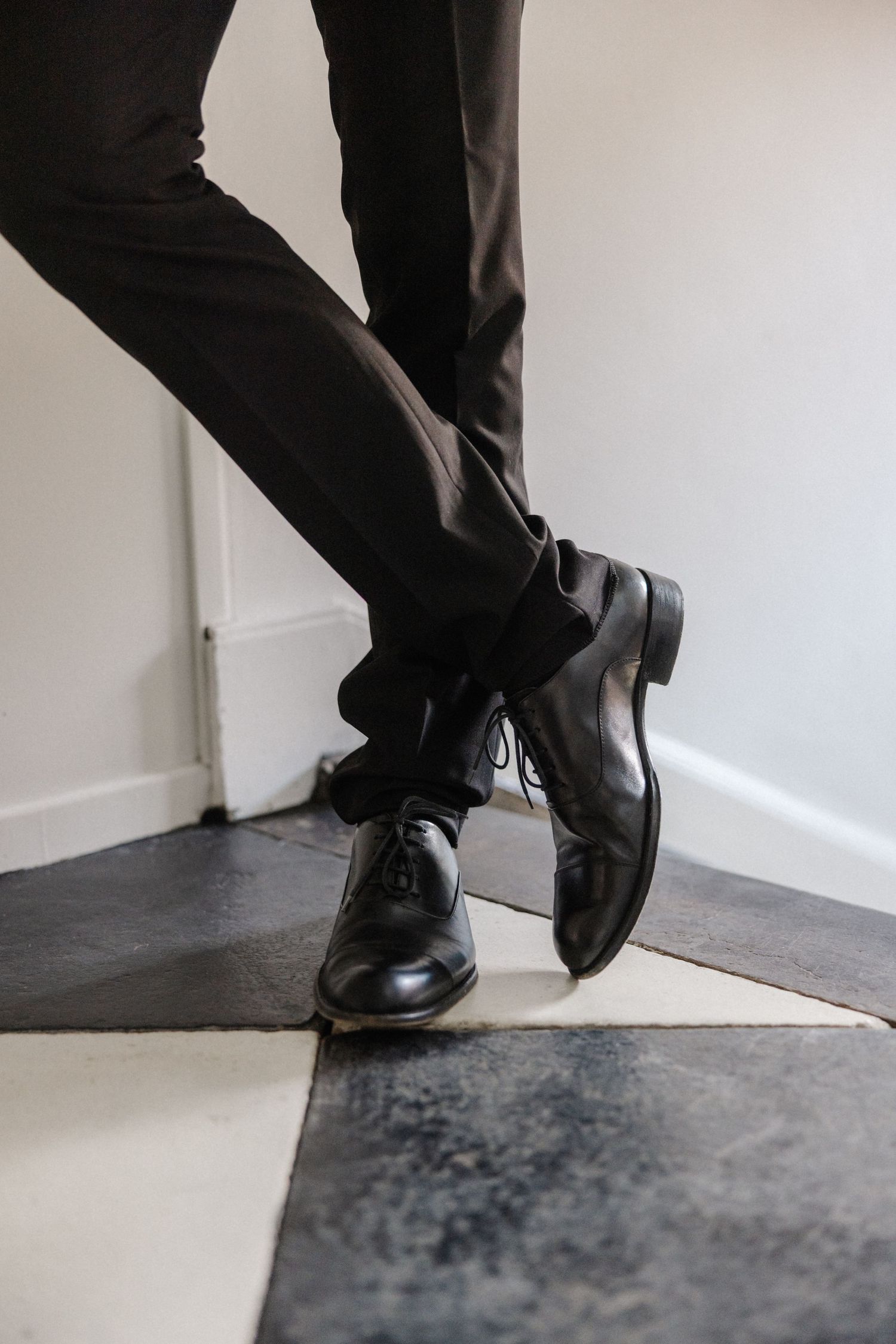 Groom putting on polished black Oxford dress shoes with dark suit trousers on checkered floor.