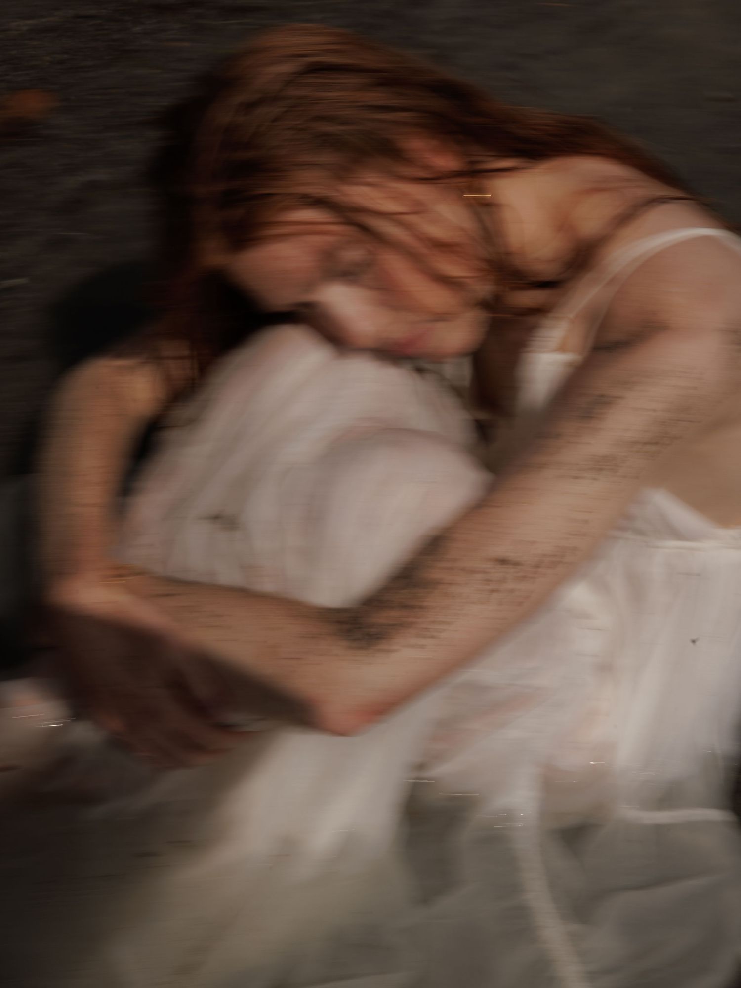 Blurred motion photograph of woman in muddy white dress curled on dark ground.