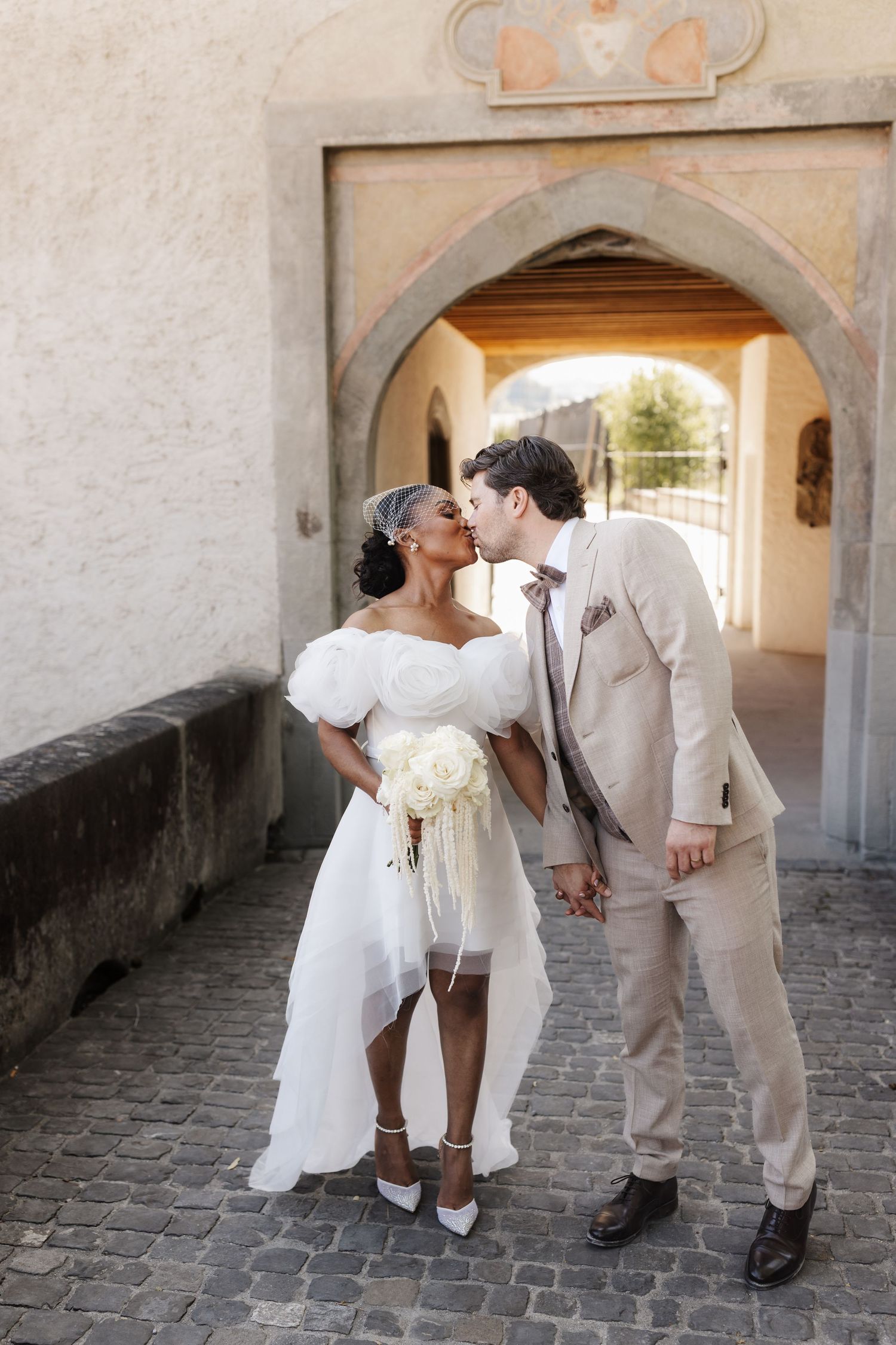 Interracial newlyweds share a kiss under stone archway, bride in white dress holding white floral bouquet.