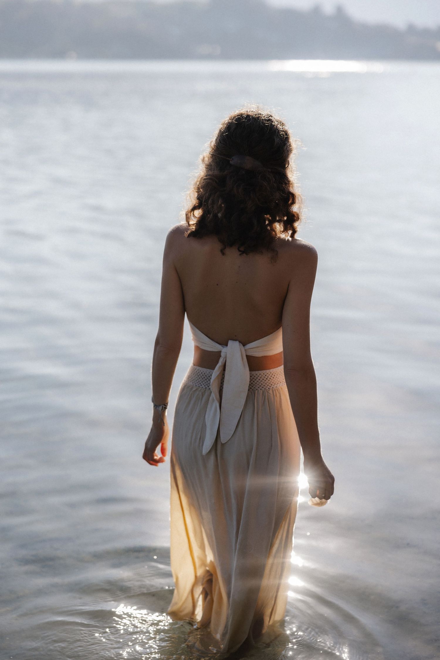Back view of woman in open-back cream halter dress standing in sunlit lake with glowing light reflection.