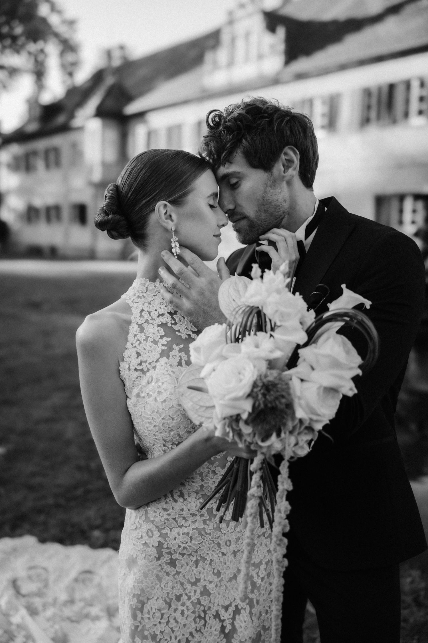 Black and white photo of groom kissing brides forehead while she holds white rose bouquet outdoors.