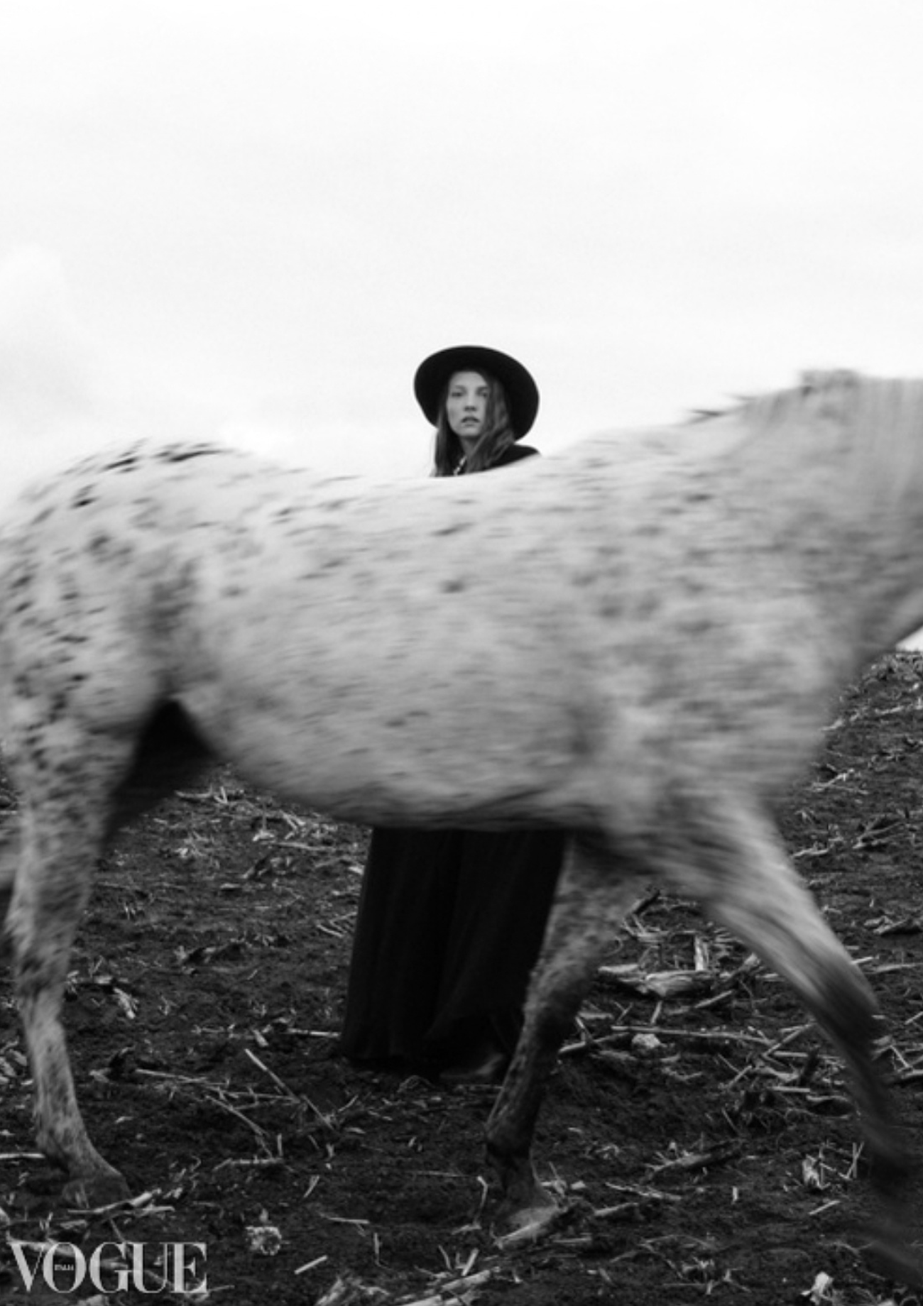 Black and white Vogue editorial of woman in hat standing beside spotted horse.